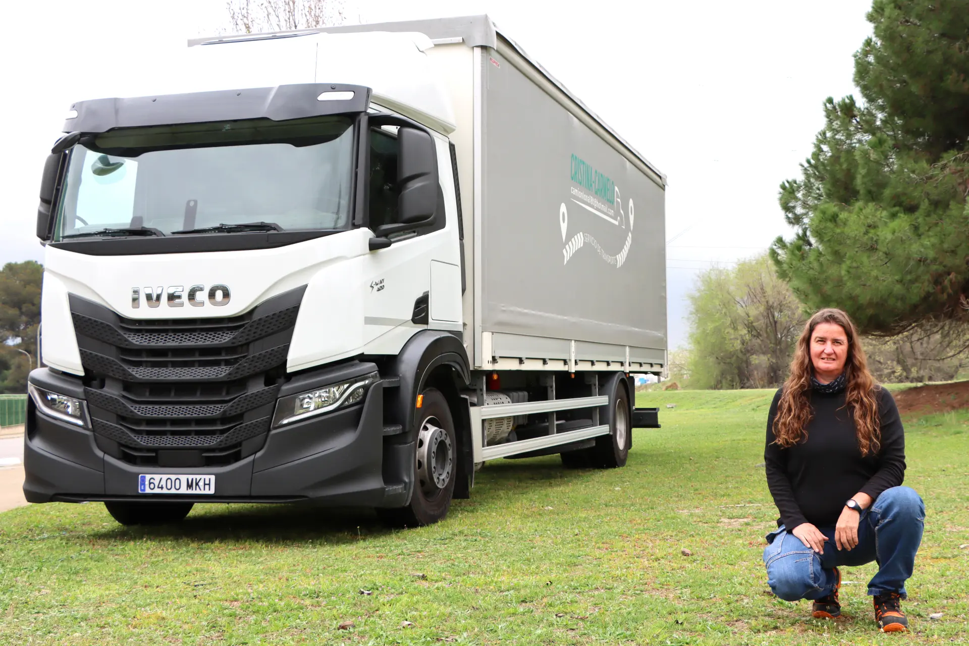 Cristina Batiste Serra se arrodilla sobre el césped junto a un camión Iveco blanco estacionado al aire libre cerca de árboles, en un momento que parece un obituario viviente a una reflexión tranquila.
