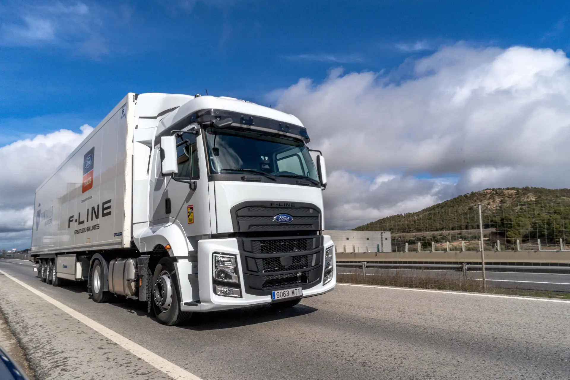 Un camión semirremolque Ford Trucks F-Line 1845T blanco circula por una carretera con colinas y nubes al fondo, ideal para quienes buscan ventajas de cambio de 16 velocidades.