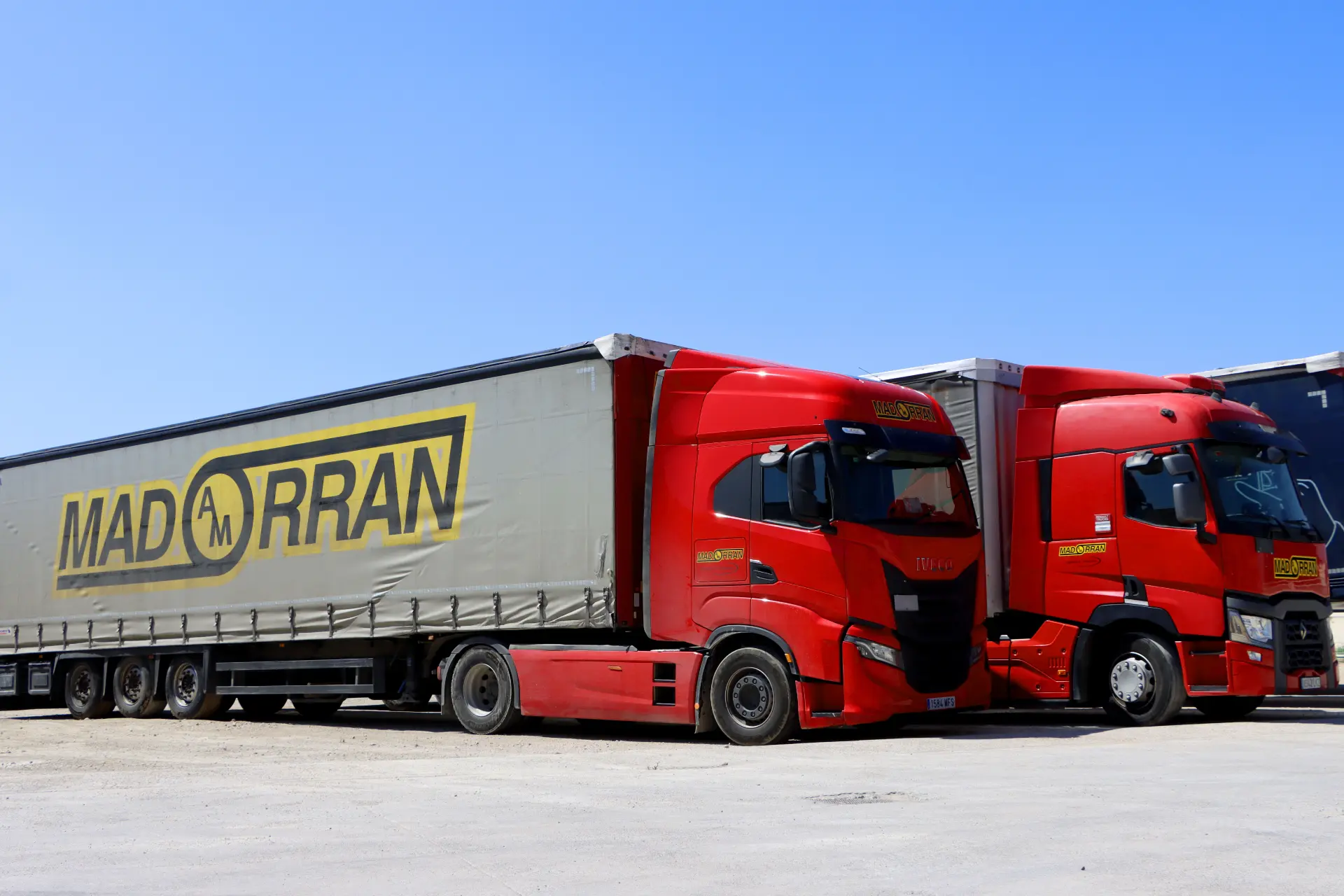 Dos semirremolques rojos con "MADRAN" en los remolques, que cuentan con un sistema Borrador automático, están estacionados uno al lado del otro bajo un cielo azul claro.