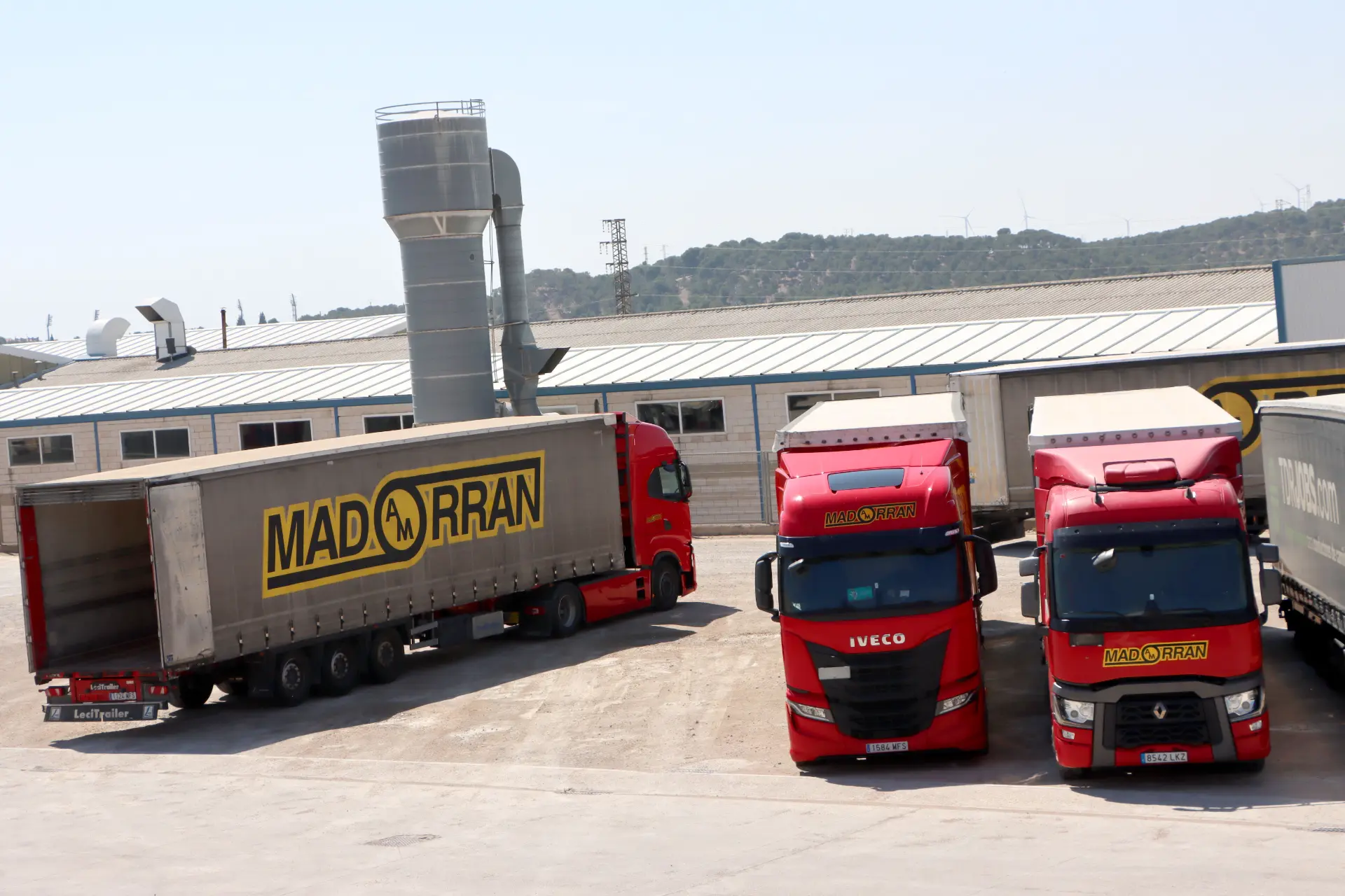 Tres camiones rojos MADRAN, equipados con sistemas Borrador automático, están aparcados junto a una nave industrial en un día soleado.