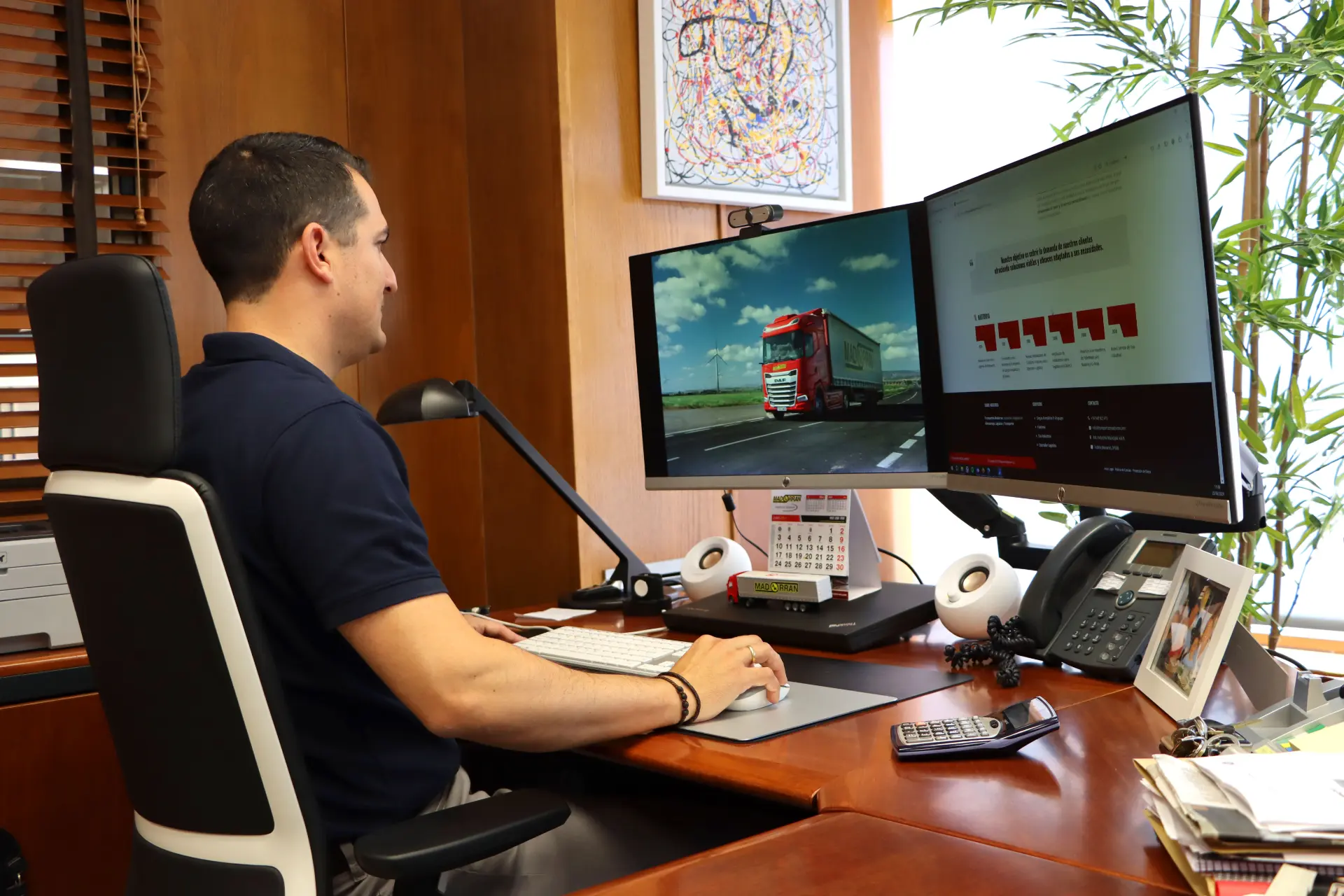 Hombre en una oficina trabajando en un escritorio con dos monitores grandes, uno mostrando un camión y el otro un gráfico rotulado "Borrador automático".