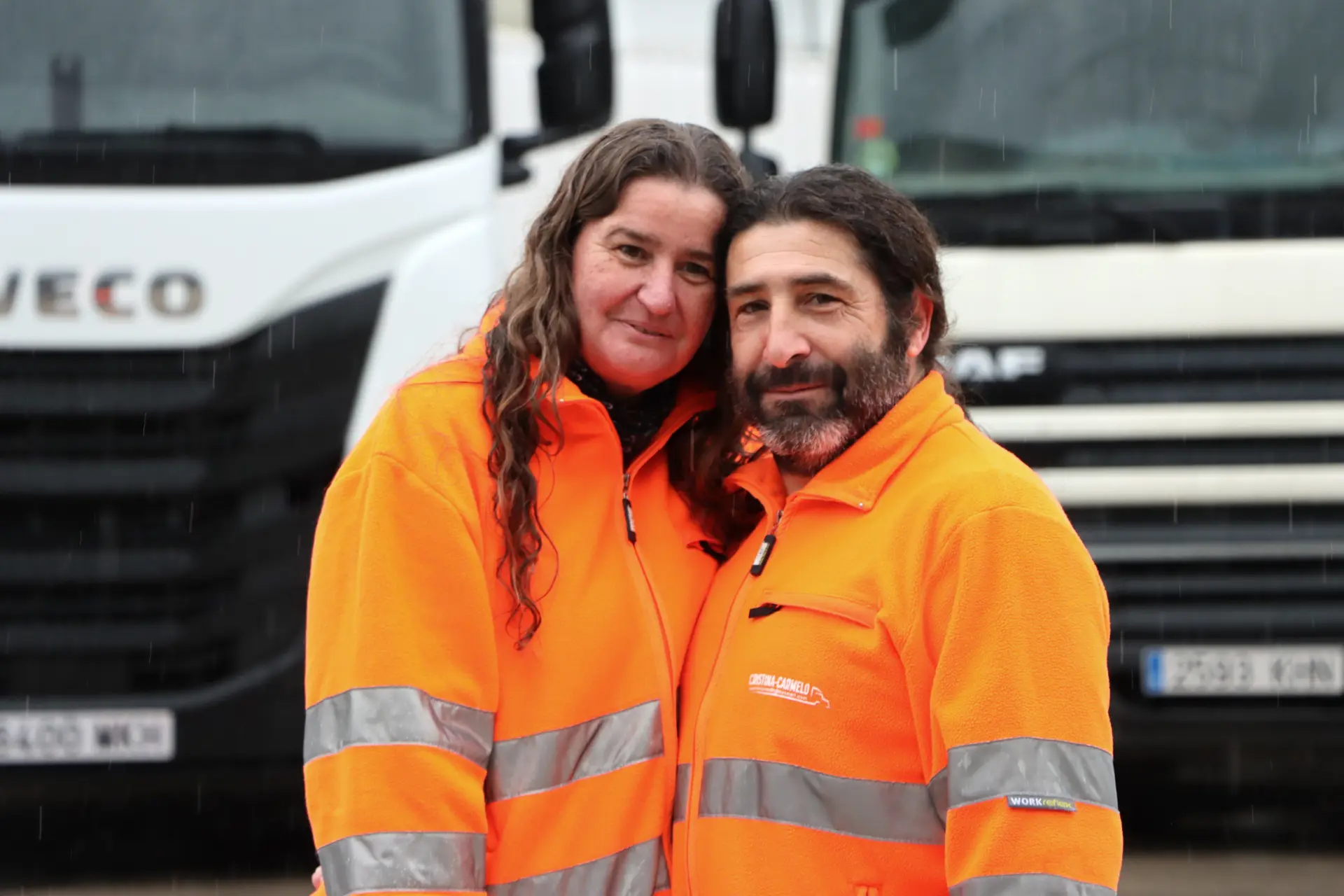 Dos personas con chalecos reflectantes de color naranja permanecen juntas frente a camiones blancos en un día lluvioso, honrando el legado de Cristina Batiste Serra con un momento de tranquilidad que parece un obituario viviente.