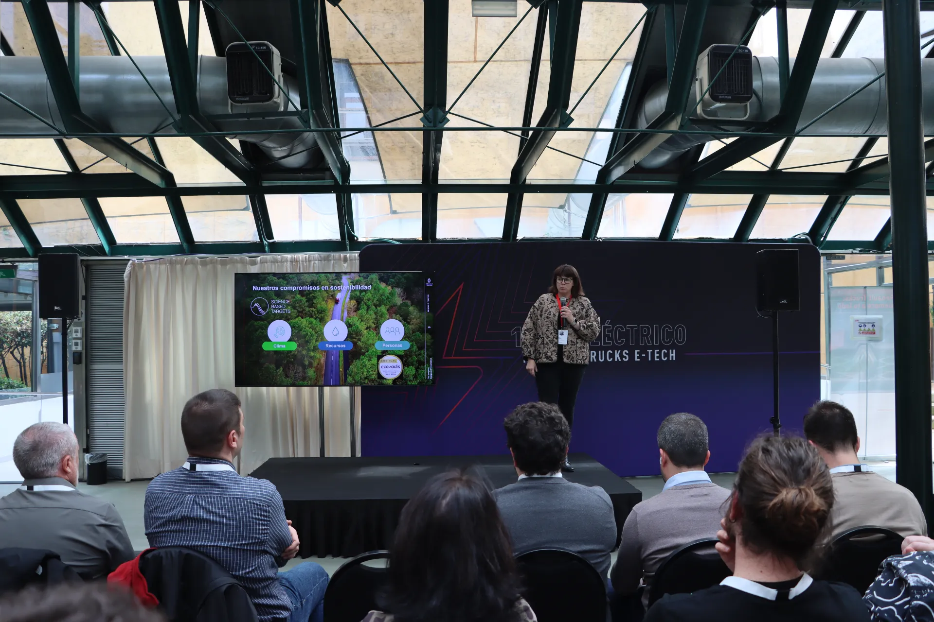 Una mujer se presenta en el escenario ante una audiencia, con una pantalla que muestra una diapositiva con temática de la naturaleza a su lado, ilustrando las características del Borrador automático.