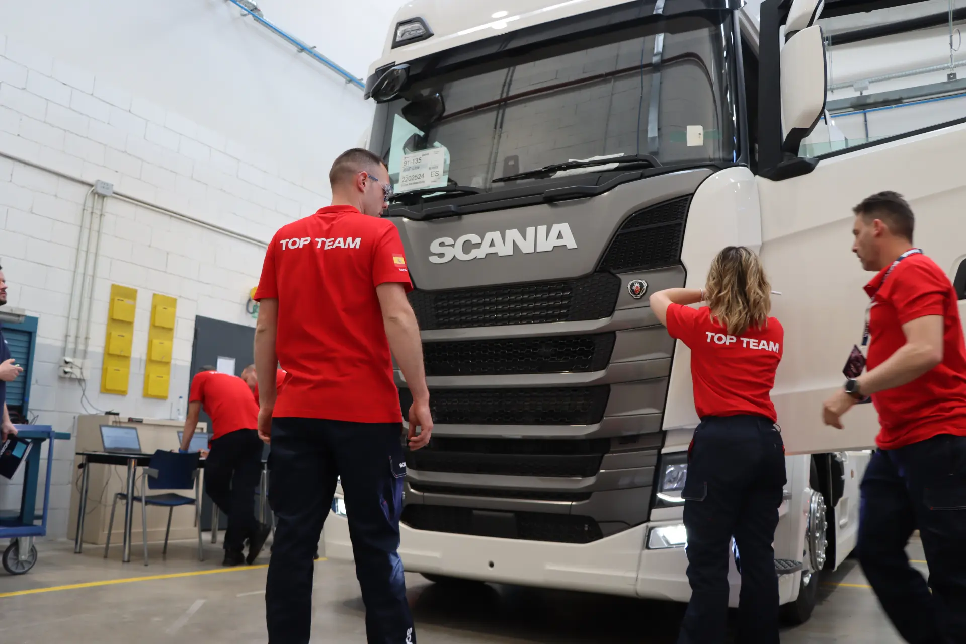 Tres personas con camisetas rojas del Scania Top Team se encuentran frente a un camión Scania blanco dentro del taller de Scalevante, listos para la competición de mecánicos.