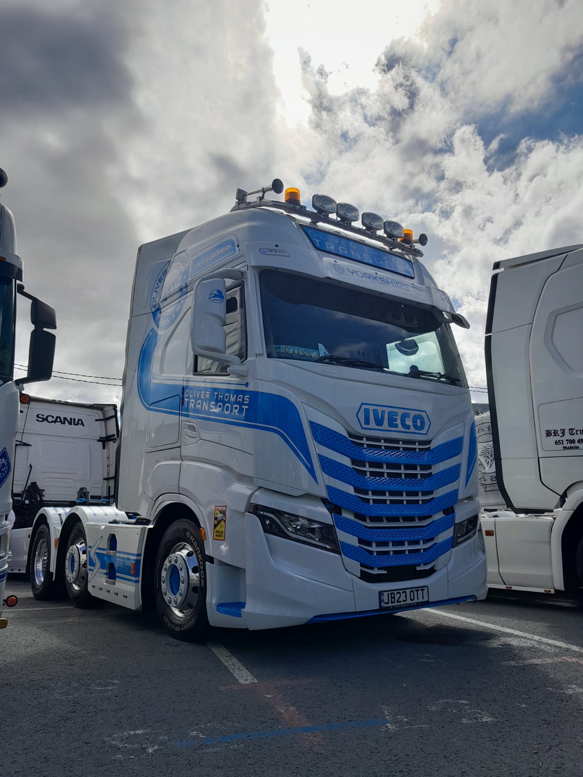 Un camión Iveco blanco con detalles en azul, que muestra la personalización de los camiones Iveco S-Way, está estacionado junto a otros camiones bajo un cielo nublado, un ajuste perfecto para las empresas de transporte familiar en Europa.