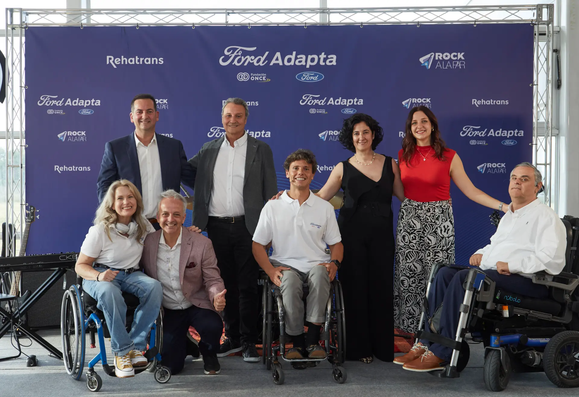 Ocho personas, algunas en sillas de ruedas, sonríen juntas frente al telón de fondo del evento Ford Adapta que muestra vehículos de adaptación rehatrans y coches para sillas de ruedas.