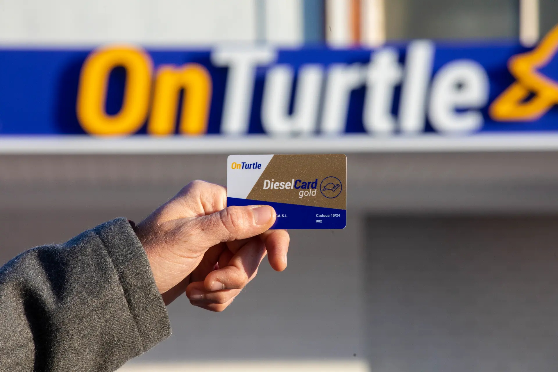 Una mano sostiene una tarjeta Diesel Gold de OnTurtle con la función de borrador automático frente a un cartel de OnTurtle.