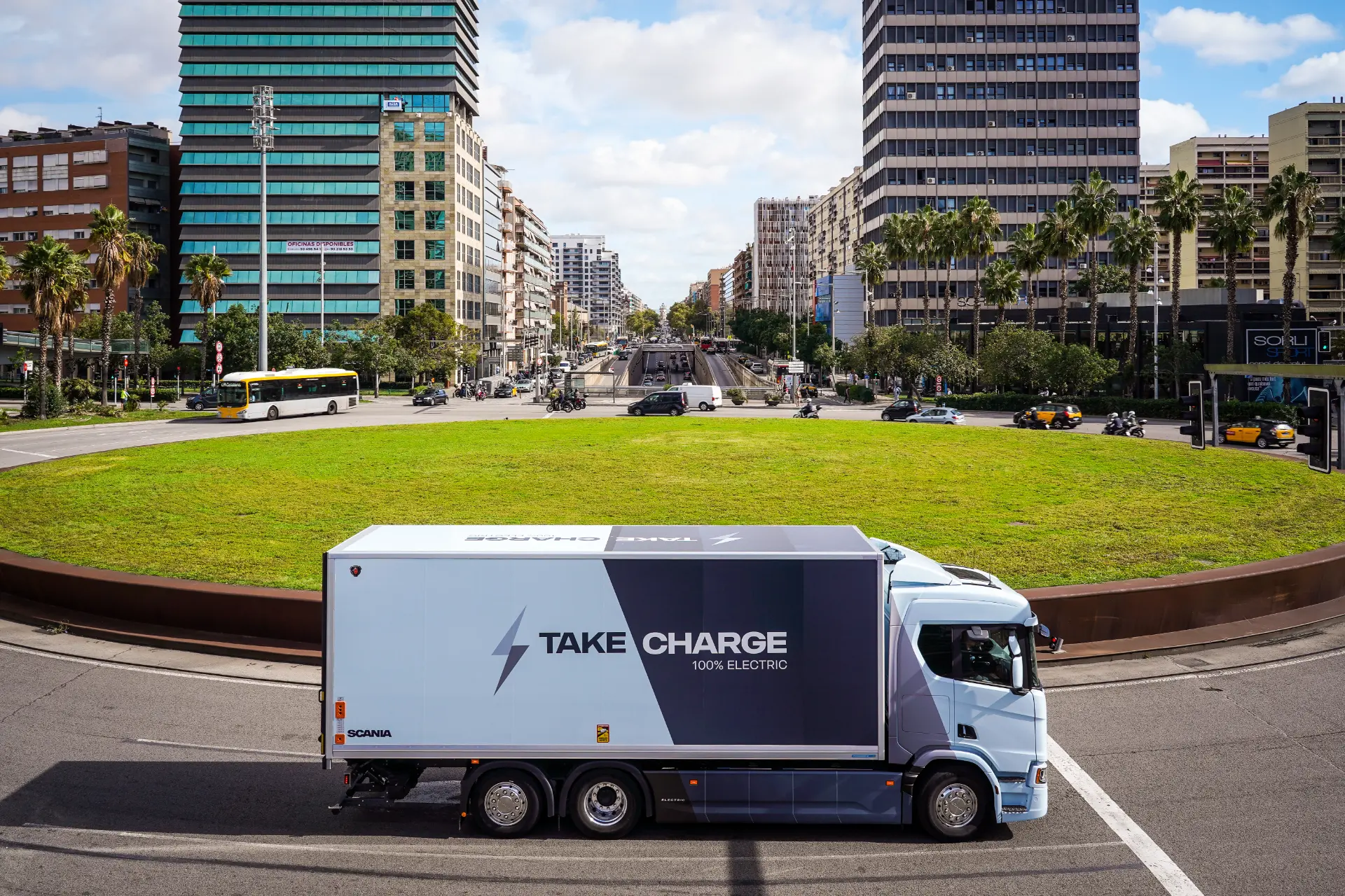 Un Scania 40R eléctrico con la marca "Take Charge" pasa por una rotonda verde en una ciudad con edificios altos, lo que resalta su desempeño en pruebas en ciudad y carretera.