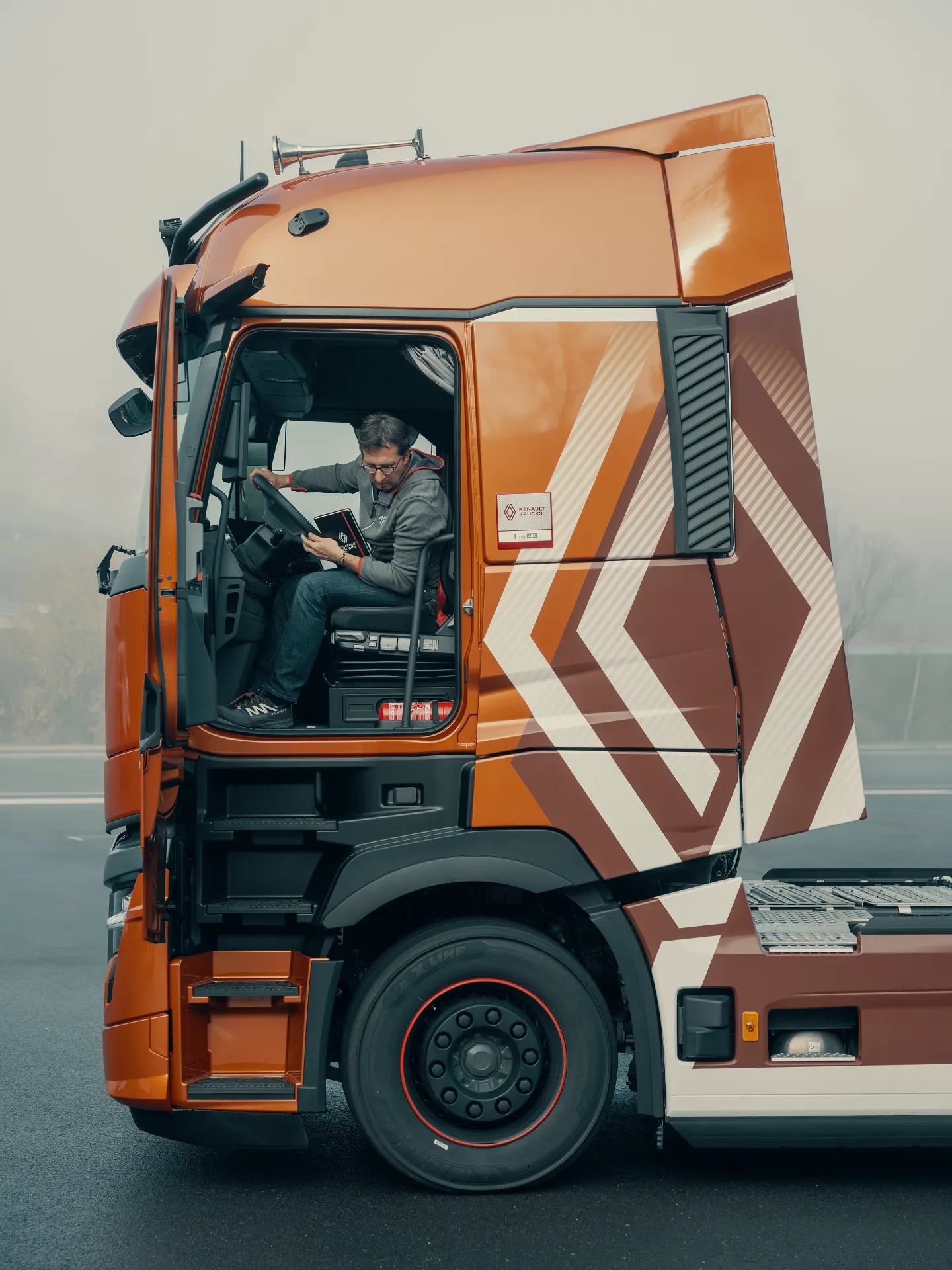 Un hombre se sienta y sonríe dentro de un gran camión naranja y blanco con la puerta de la cabina abierta en un día de niebla, su mano sosteniendo un borrador automático.