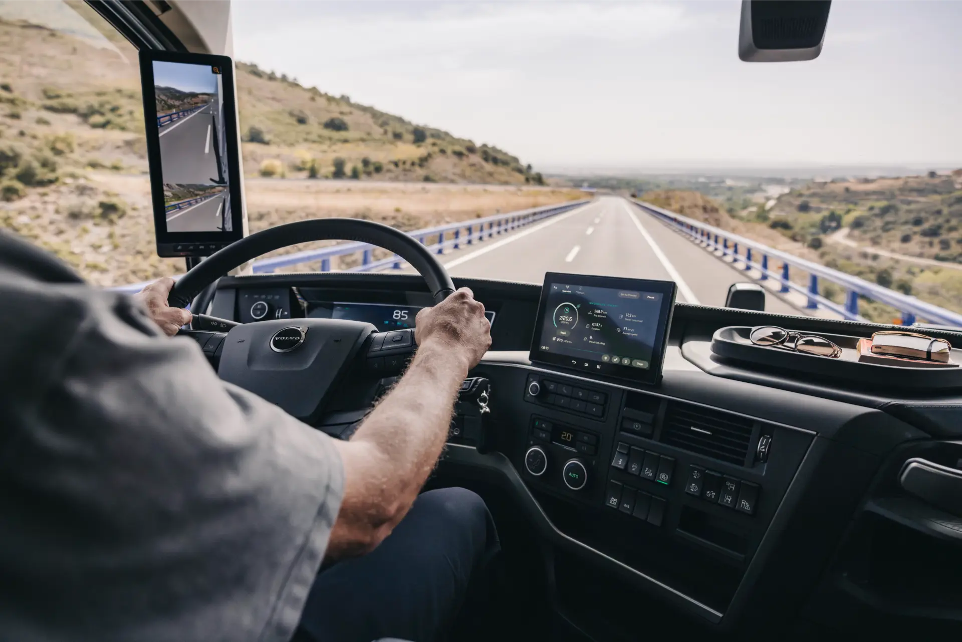 Persona que conduce un camión en una carretera, el tablero muestra pantallas digitales que muestran tecnología de camiones Volvo, mientras el camino se extiende frente a ella.