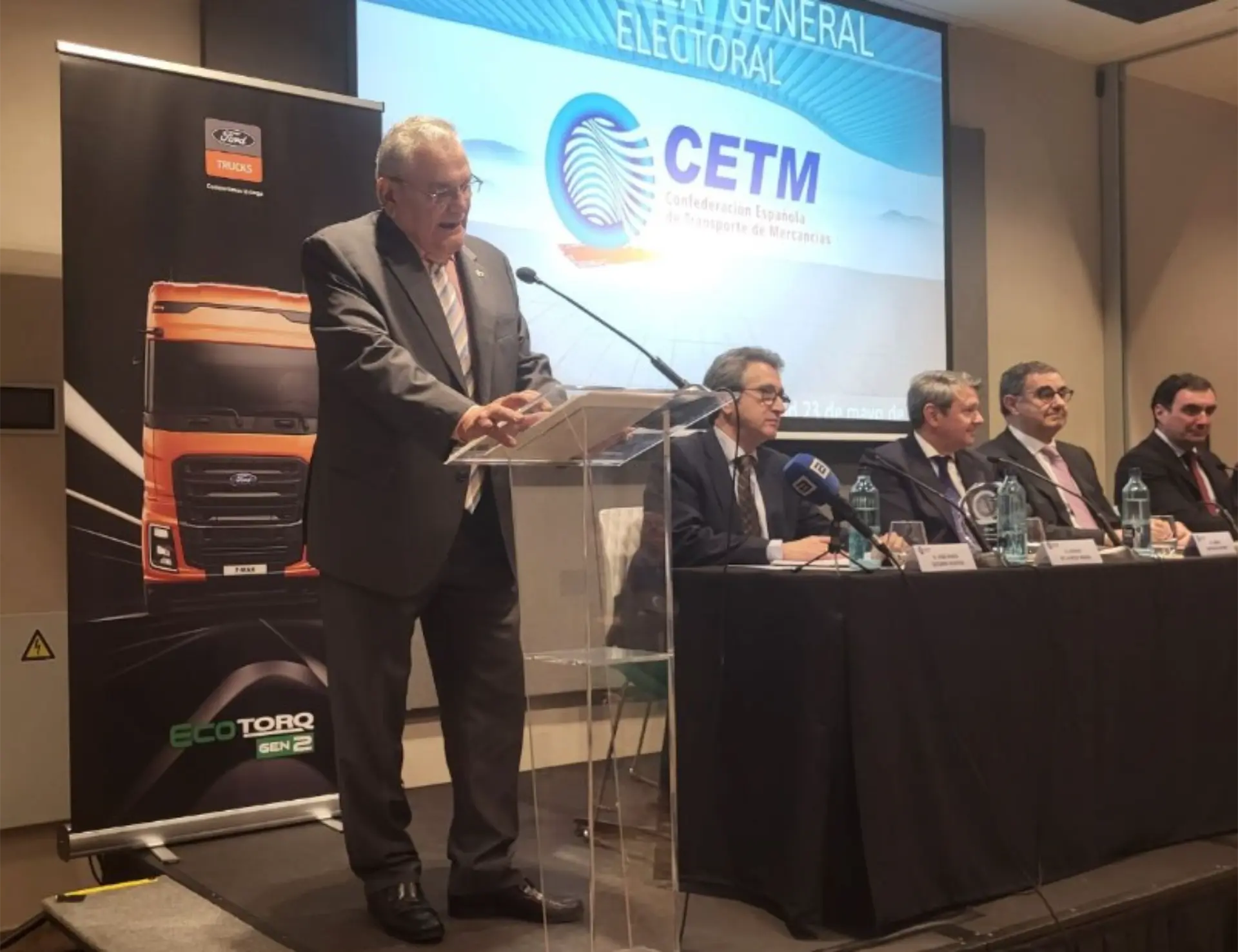 Un hombre habla en un podio durante una conferencia sobre los problemas del sector del transporte en España, con cuatro panelistas sentados y una pancarta de un camión cerca.