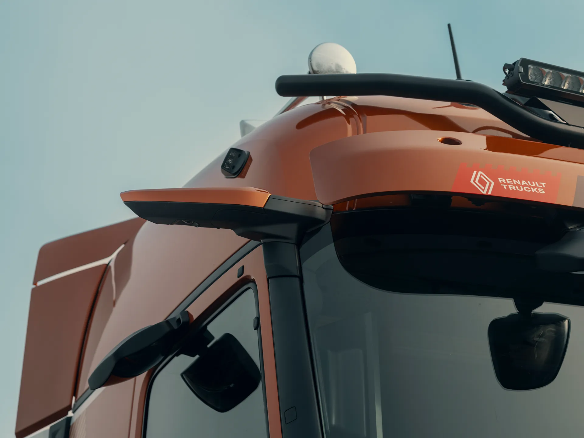 Primer plano de la cabina de un camión Renault rojo, donde se aprecia el espejo lateral, el logotipo de Renault Trucks y un vistazo al interior con un borrador automático en el salpicadero.