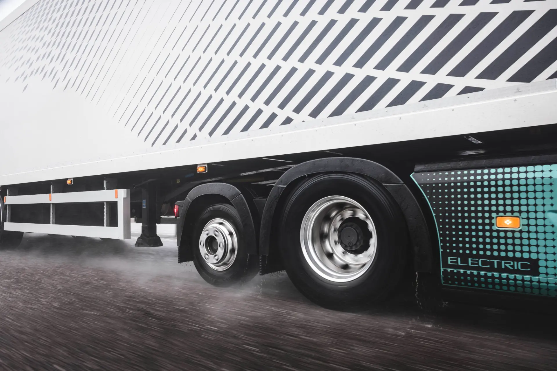 Primer plano de las ruedas de un camión eléctrico circulando por una carretera mojada, con niebla de agua detrás de los neumáticos.