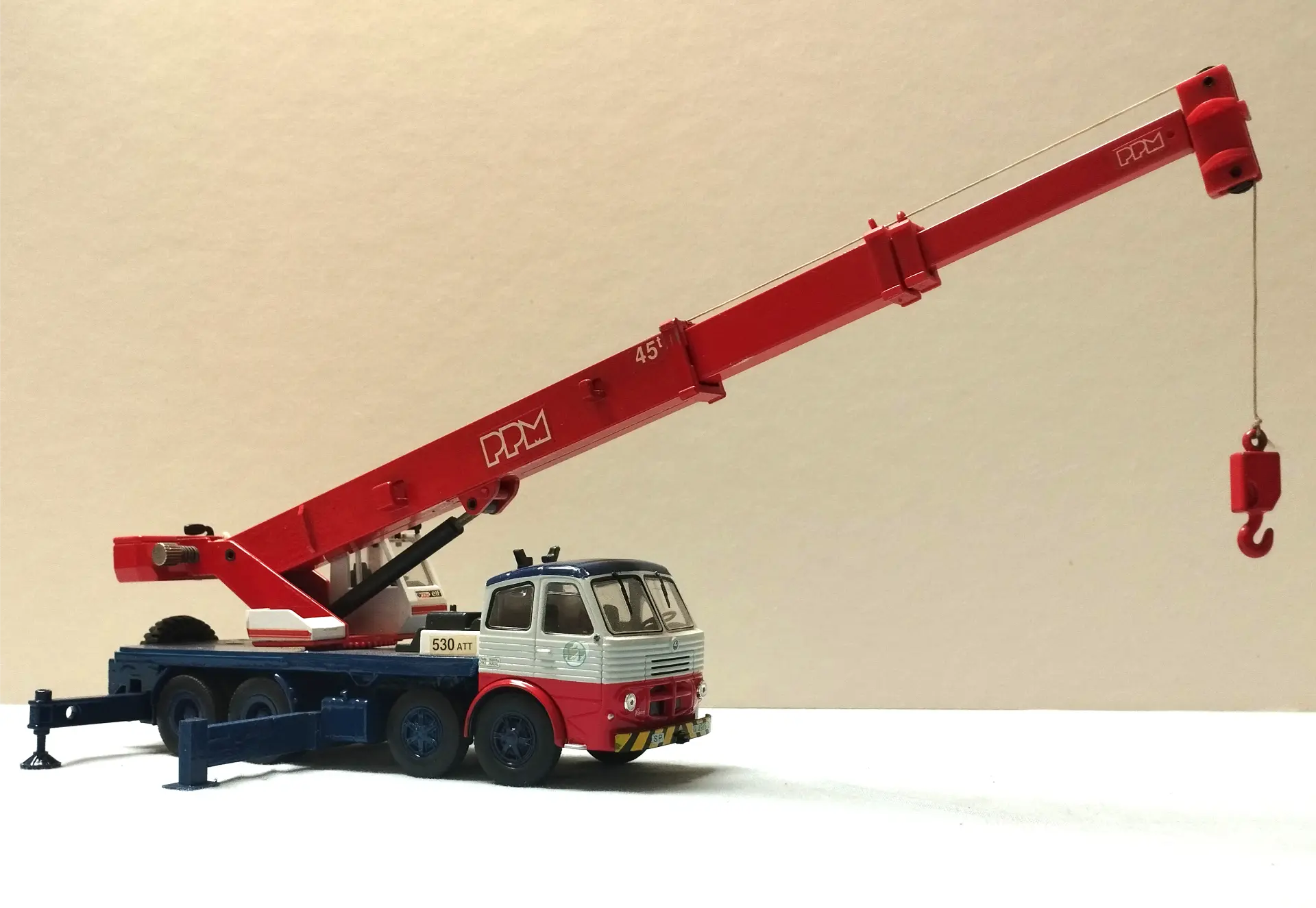 Un camión grúa de juguete rojo y blanco con una pluma extendida y un gancho sobre un fondo liso, perfecto para modelismo de camiones o para agregar a su colección de maquetas de camiones a escala 1/43.