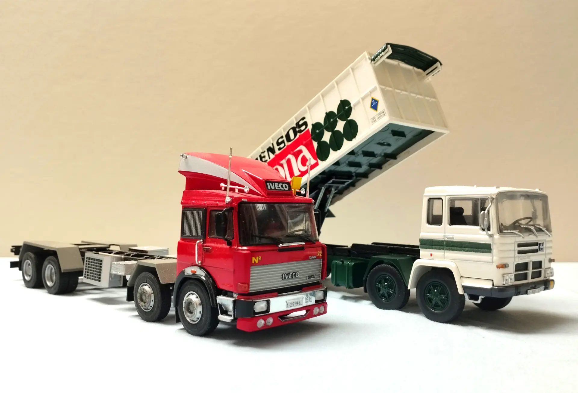 Dos modelos de camiones, uno rojo y otro blanco, con el remolque del camión blanco elevado en posición basculante, perfecto para los fanáticos del modelismo de camiones y las maquetas personalizadas.