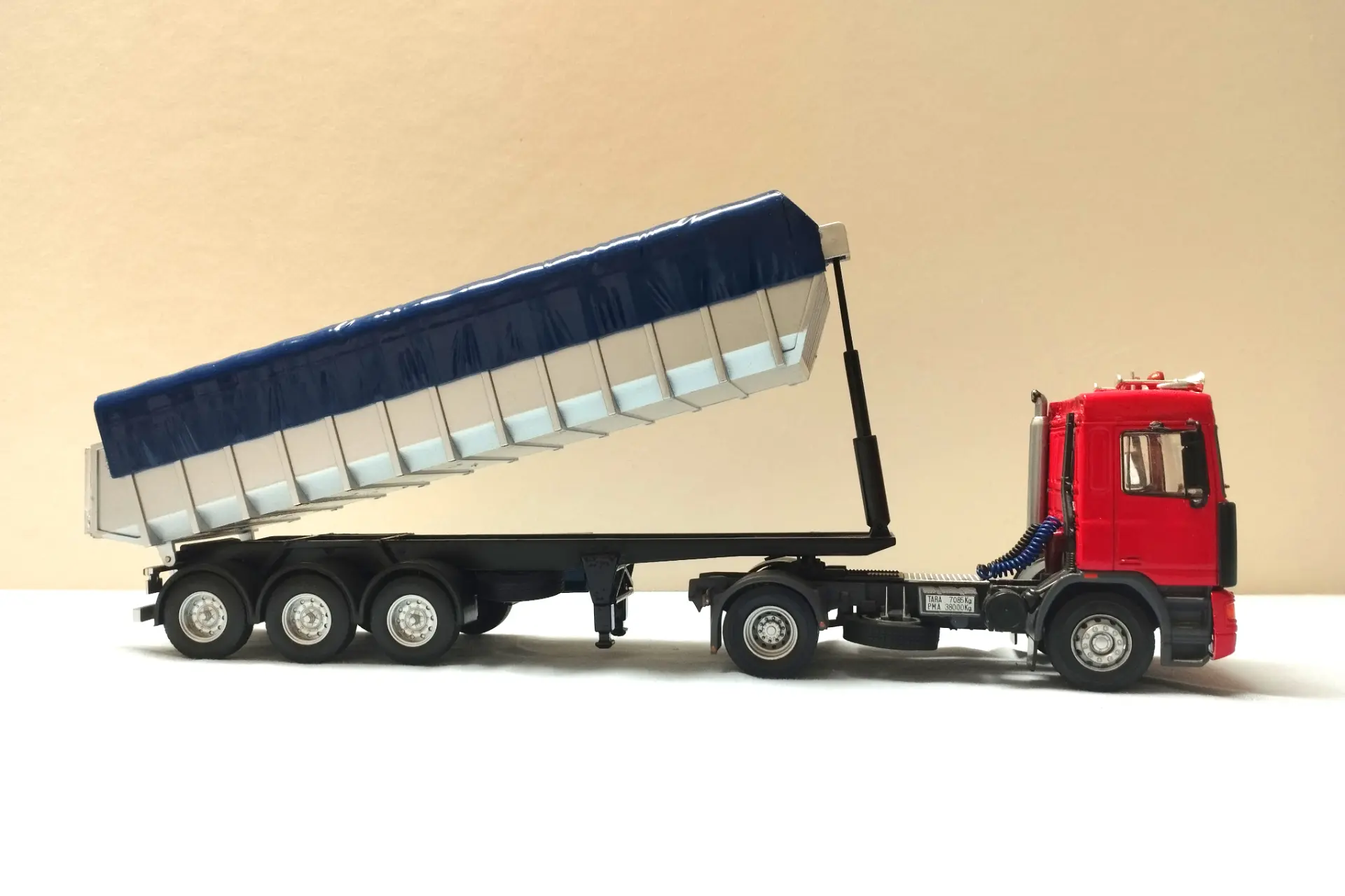Un camión volquete rojo con su caja de remolque azul y plateada levantada, mostrado en una vista lateral, perfecto para coleccionistas de maquetas de camiones o entusiastas de camiones a escala 1:43.