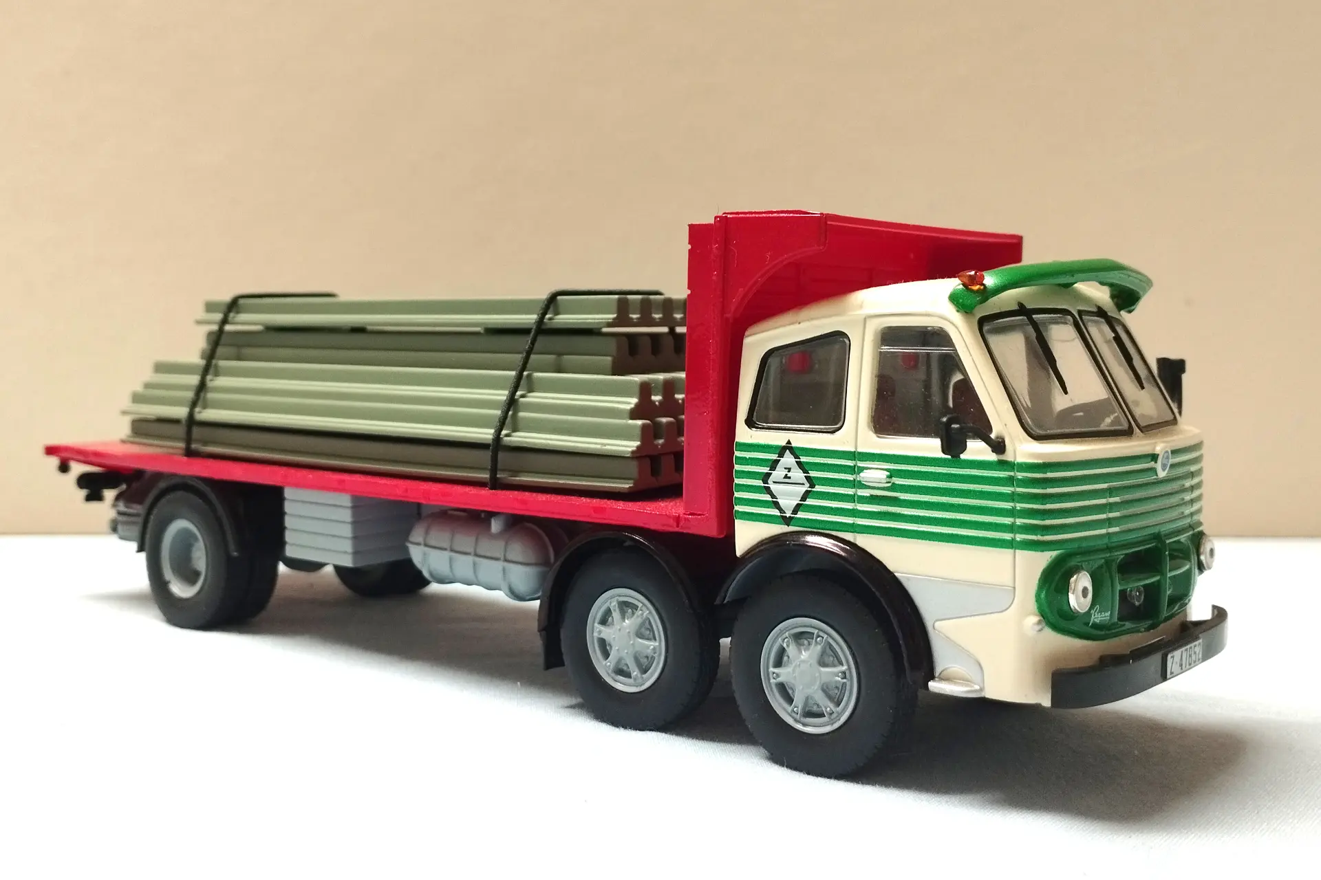 Camión de plataforma verde y blanco que transporta vigas de metal apiladas, visto en ángulo sobre un fondo liso, perfecto para modelismo de camiones o para cualquiera que busque maquetas de camiones a escala 1/43.