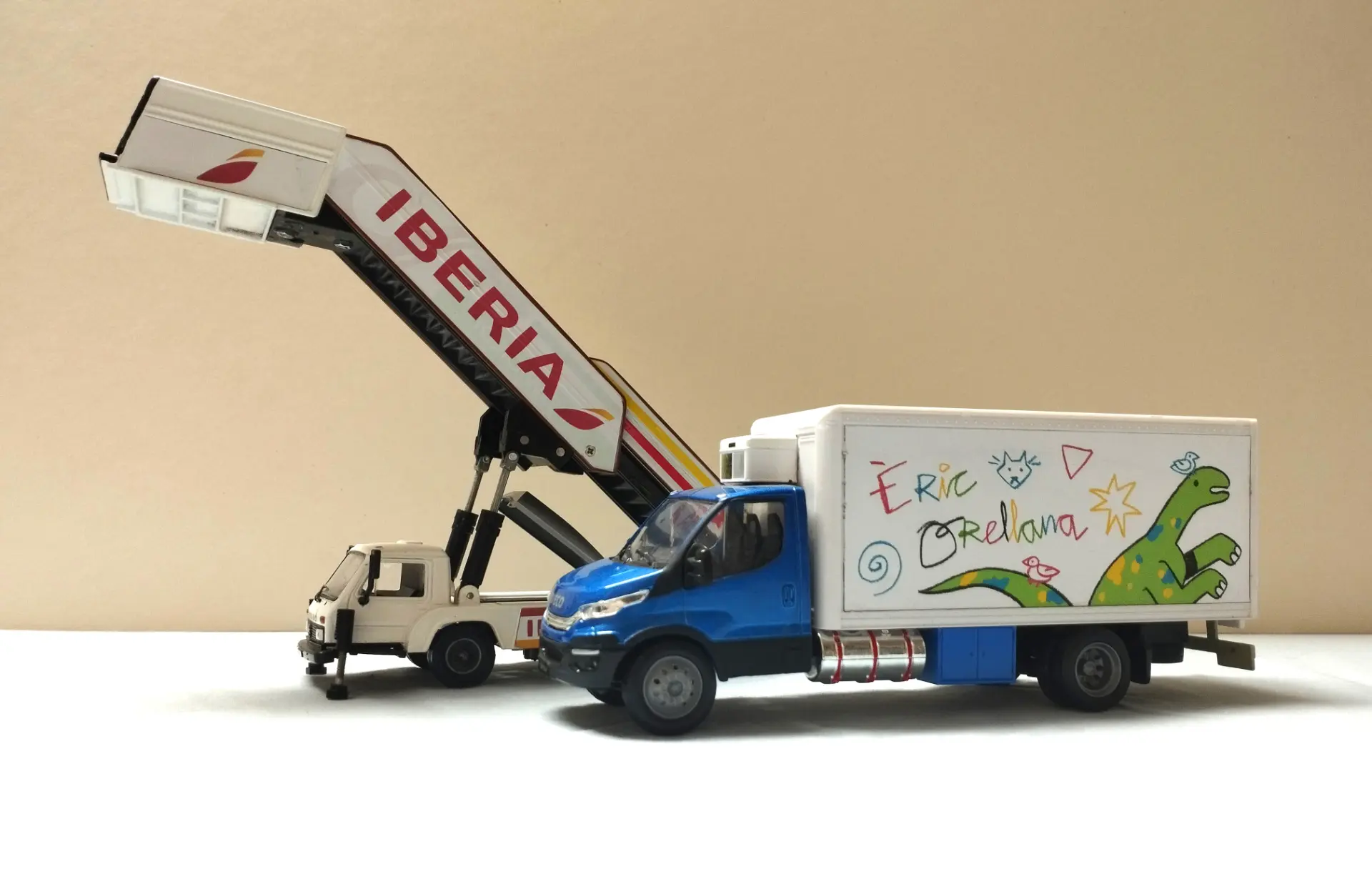Dos camiones de juguete: un camión escalera de avión de Iberia y un camión azul con un dinosaurio y garabatos en su lateral, ambos perfectos como maquetas de camiones o para coleccionistas de camiones a escala 1:43.