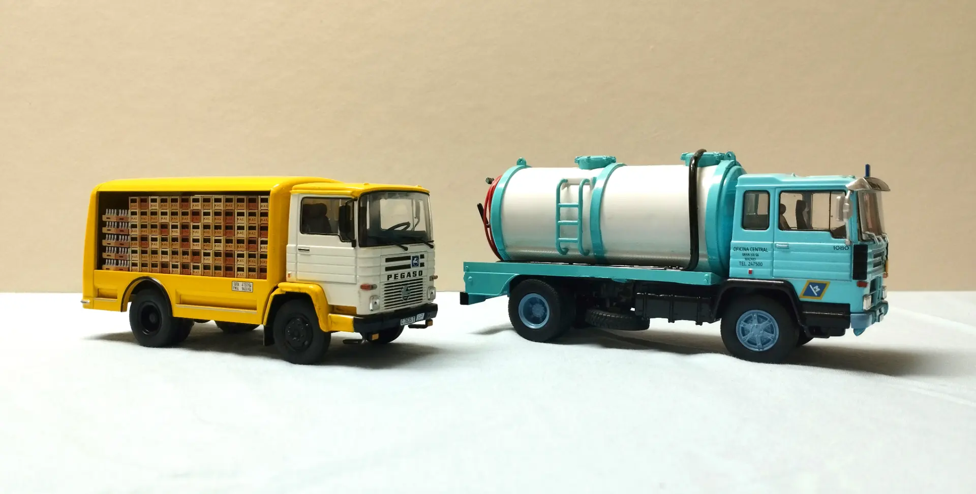 Dos camiones de juguete, uno amarillo y otro azul con un tanque blanco, se exhiben sobre una superficie blanca lisa, perfectos como maquetas de camiones o para coleccionistas de camiones a escala 1:43.