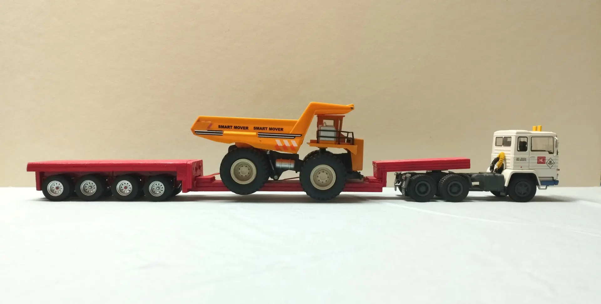 Camión de juguete y remolque que transporta un camión volquete de juguete sobre una superficie clara con un fondo beige liso, perfecto para coleccionistas de camiones a escala 1:43 o interesados en maquetas de camiones.