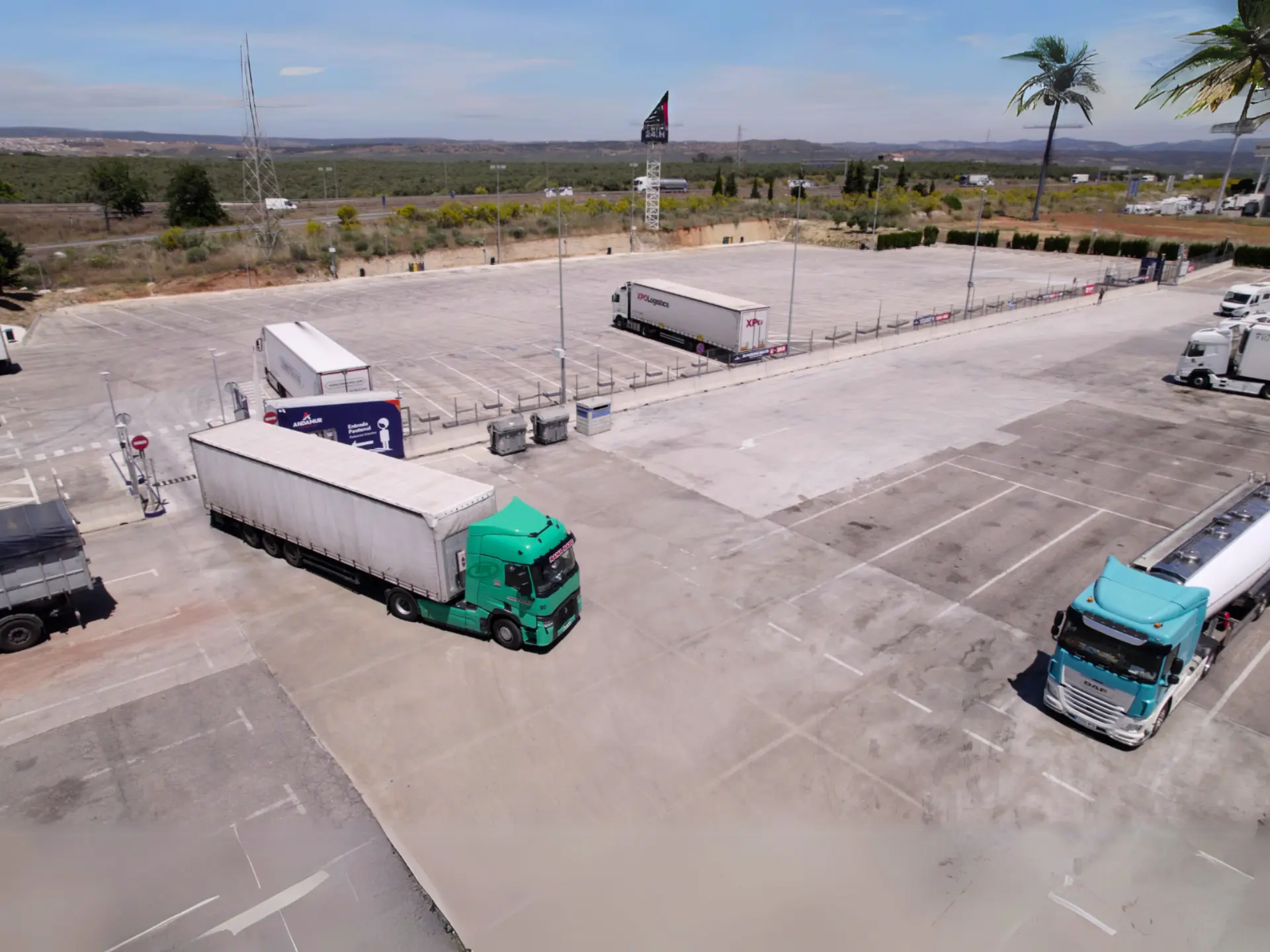 Varios camiones aparcan en el soleado aparcamiento de andamur guarromán, conocido por aparcamientos seguros transportistas, con algunos árboles y un paisaje lejano.