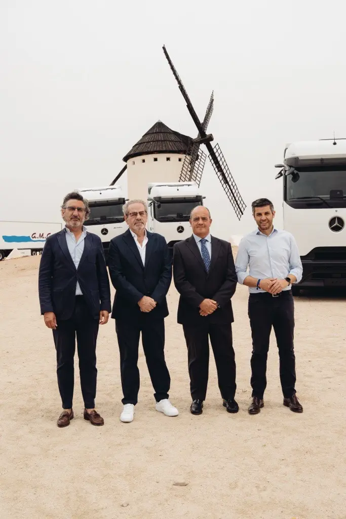 Cuatro hombres trajeados, de pie frente a dos camiones y un molino de viento en un día arenoso y nublado, discuten sobre el nuevo sistema de borrador automático.