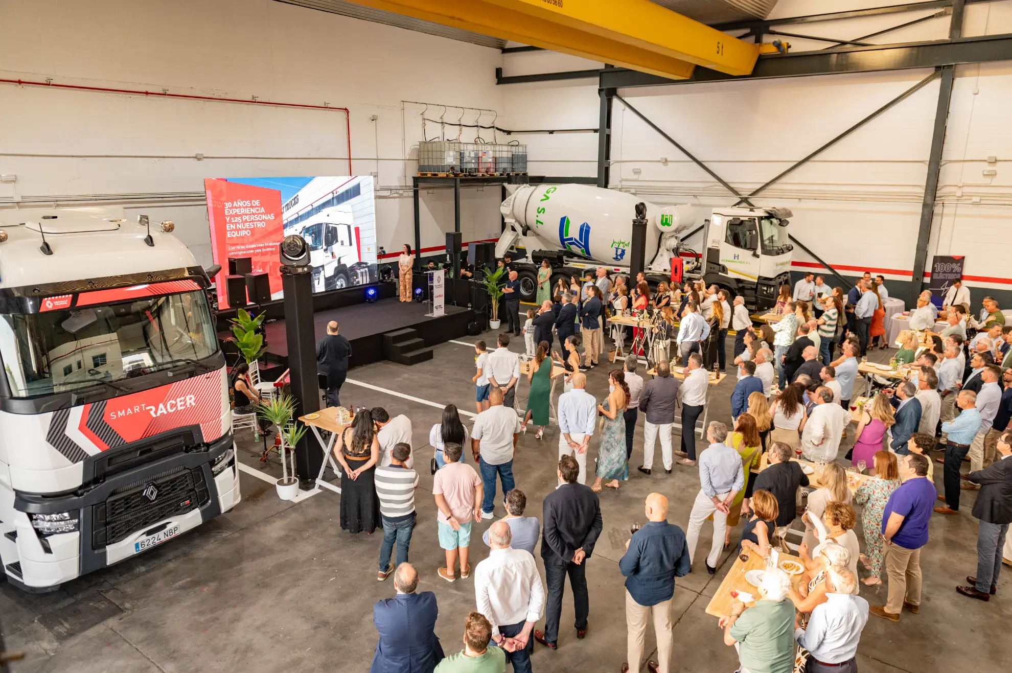Una multitud asiste a una presentación en una nave con camiones y una gran pantalla en la que se muestra un evento empresarial del distribuidor oficial camiones Andalucía, destacando los camiones eléctricos Renault Trucks.
