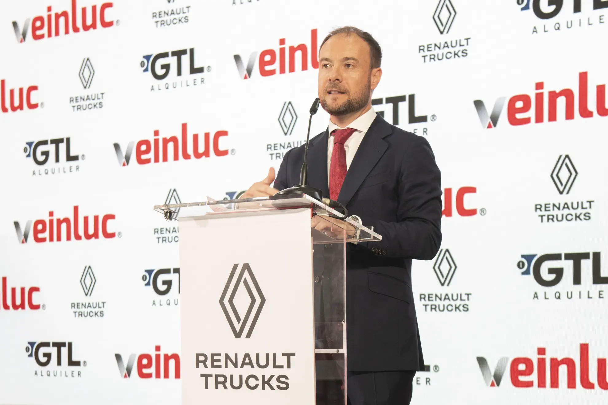 Un hombre con traje habla en un podio con Renault Trucks y otros logotipos detrás de él, destacando innovaciones como los camiones eléctricos Renault Trucks.