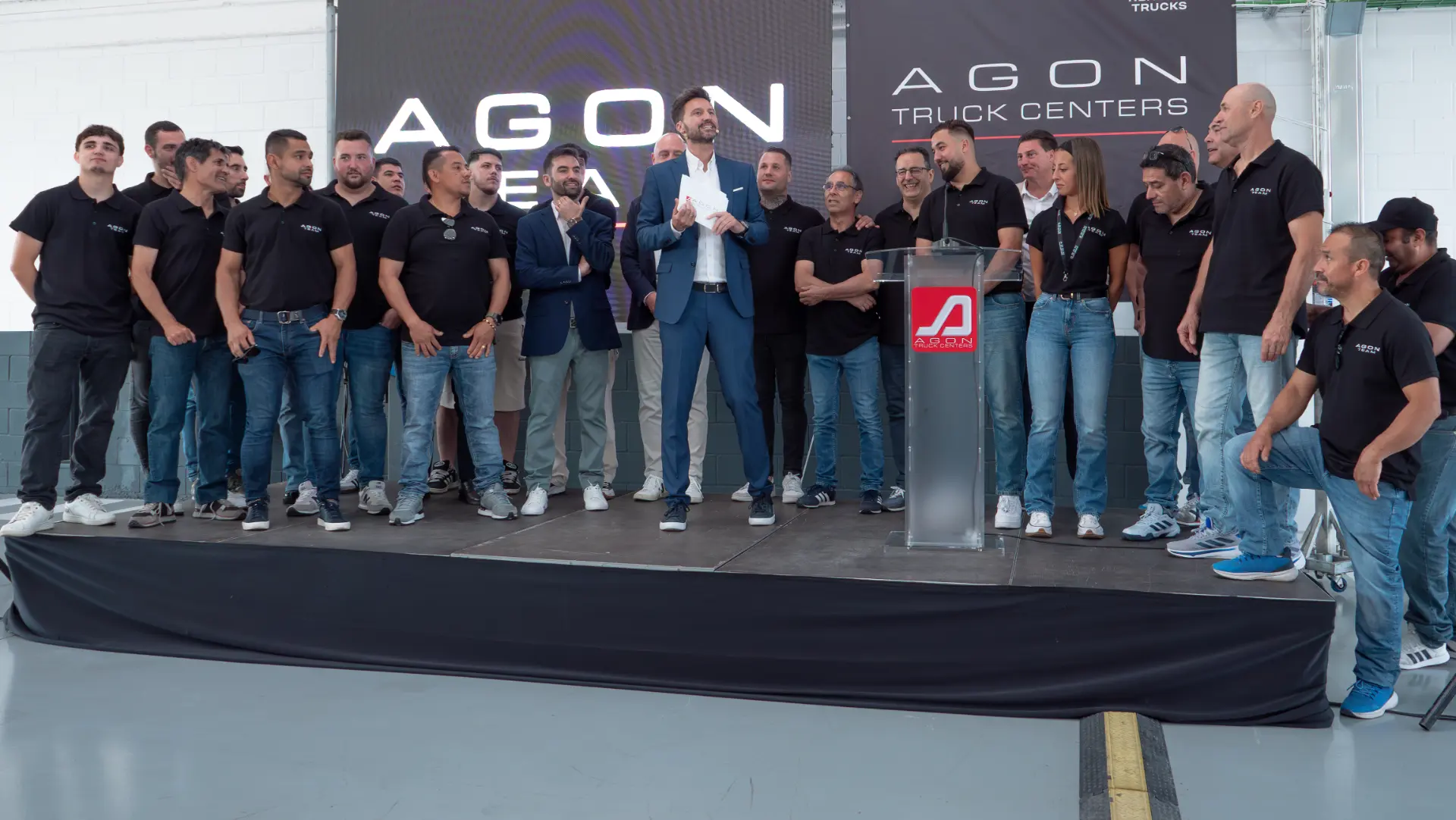 Un grupo de personas con camisas negras se encuentra en el escenario en un evento de AGON Truck Centers en Tarragona, representando al concesionario oficial Renault Trucks, con un hombre hablando en un podio.