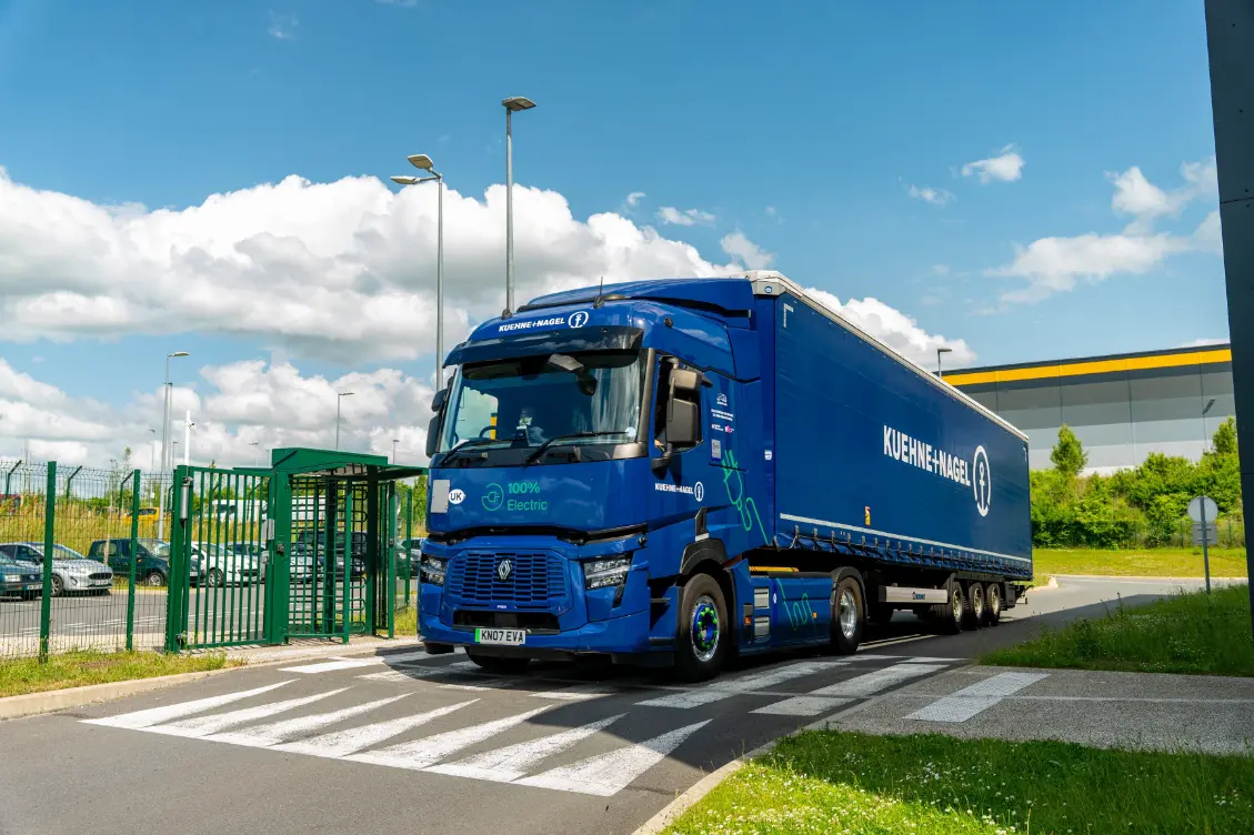Un camión eléctrico azul de Kuehne+Nagel con tecnología de Borrador automático atraviesa un paso de cebra cerca de una nave industrial en un día soleado.