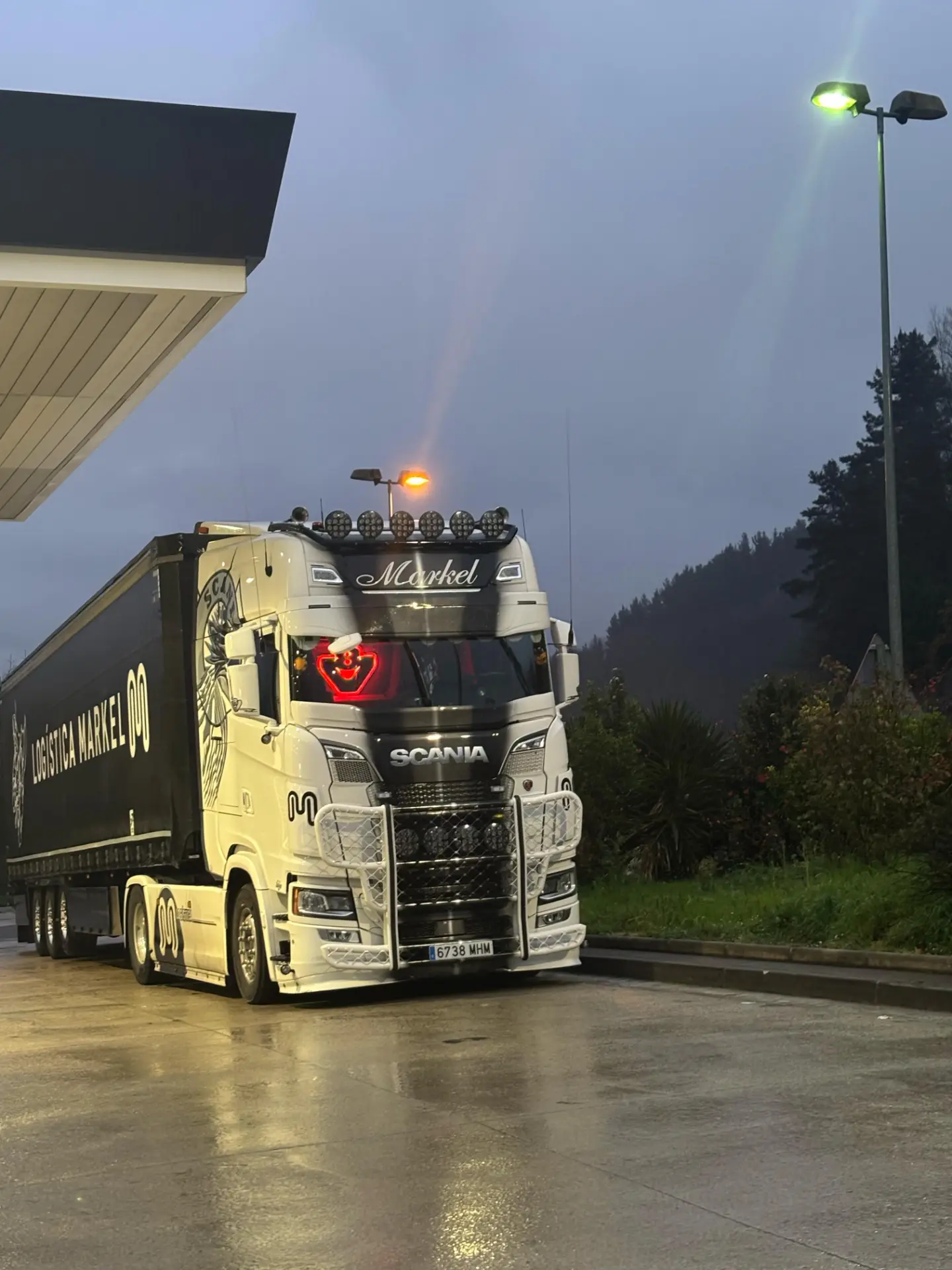 Un camión Scania blanco con remolque, marcado Solo Camión 395, está estacionado en una gasolinera en una tarde lluviosa, sus luces y el logotipo de JDC se reflejan en el suelo mojado.