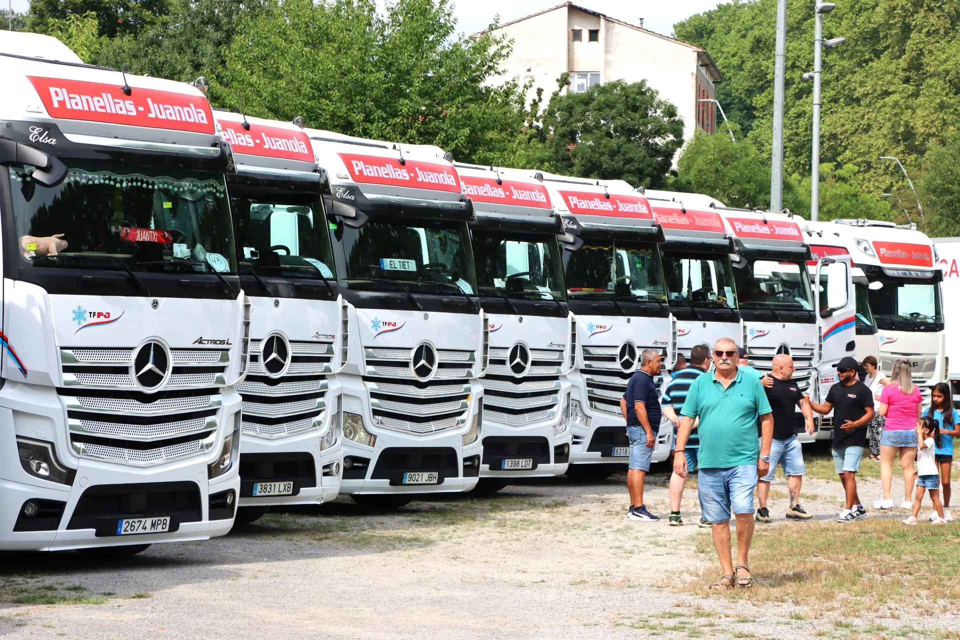 Varios camiones Mercedes blancos, que forman parte de una concentración de camiones de Olot, están aparcados en fila mientras la gente camina y se para cerca, al aire libre.