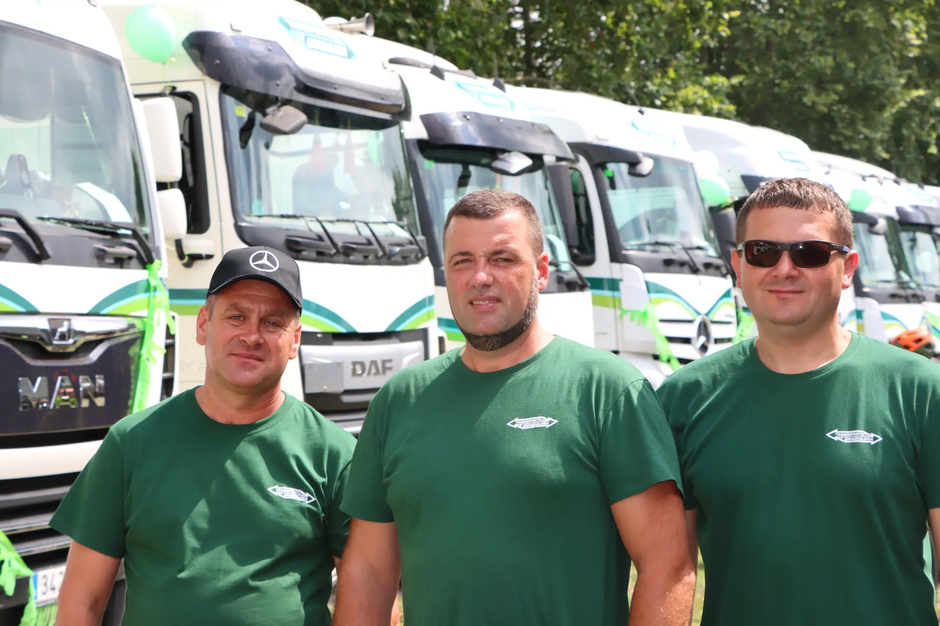 Tres hombres con camisas verdes frente a una fila de camiones de Olot en un día soleado.