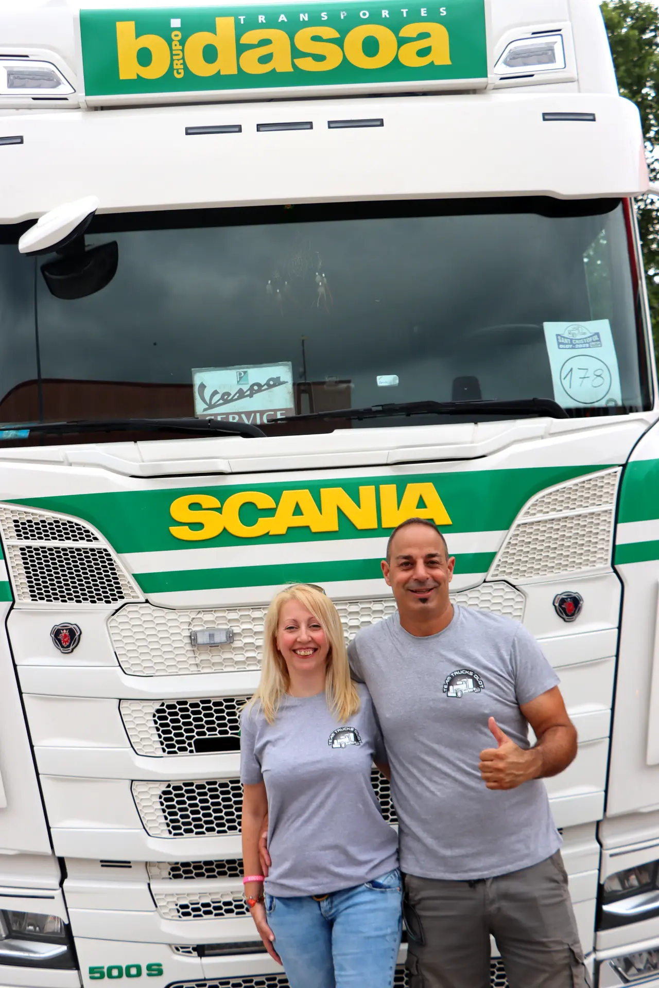 Dos personas sonriendo y posando delante de un camión Scania blanco con rótulos verdes y amarillos en la XI Concentración, un evento destacado de camiones de Olot.