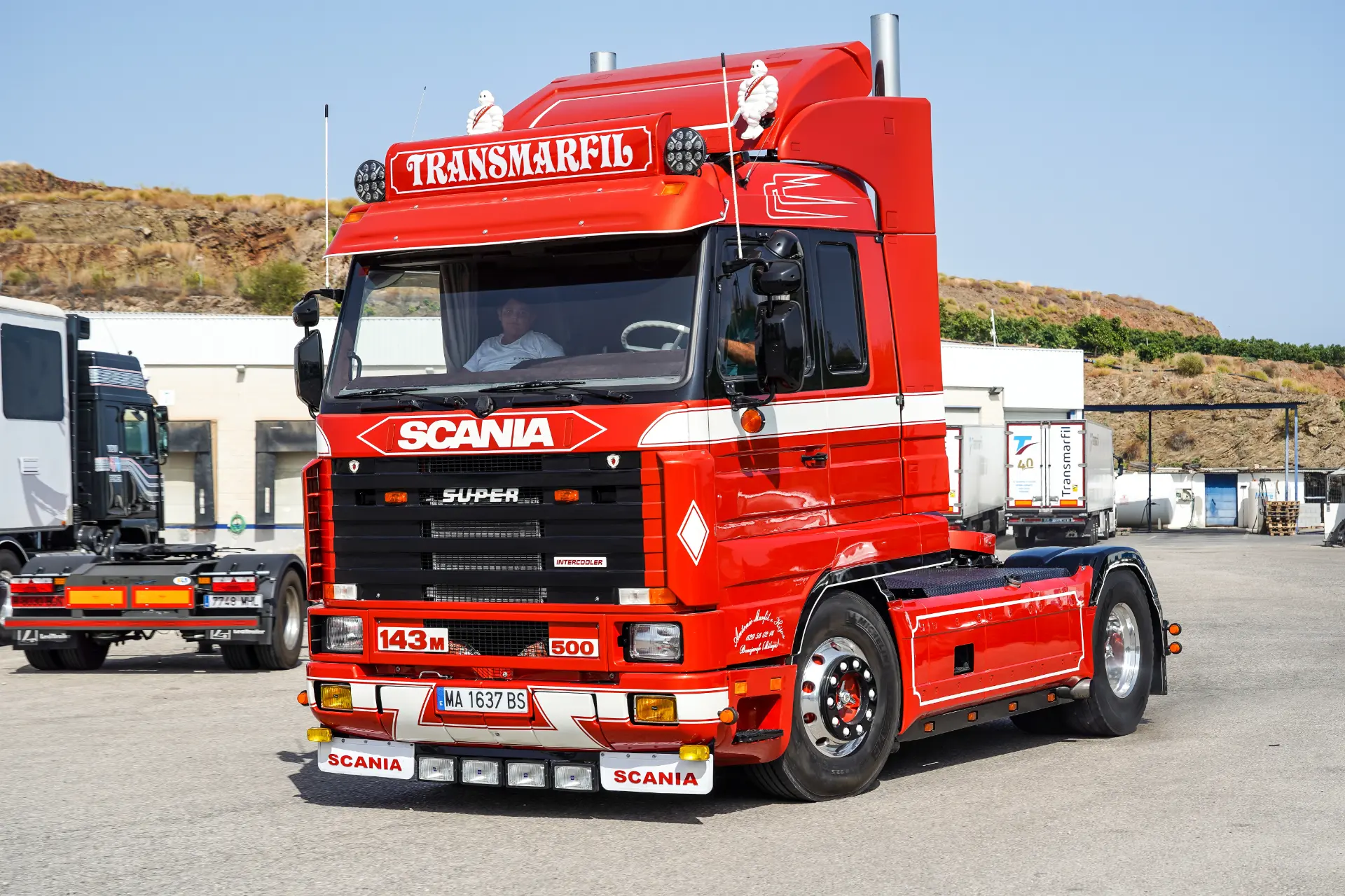 Camión Scania rojo de época rotulado "Transmarfil" con una pegatina de Borrador automático aparcado en una zona industrial, personas en su interior.