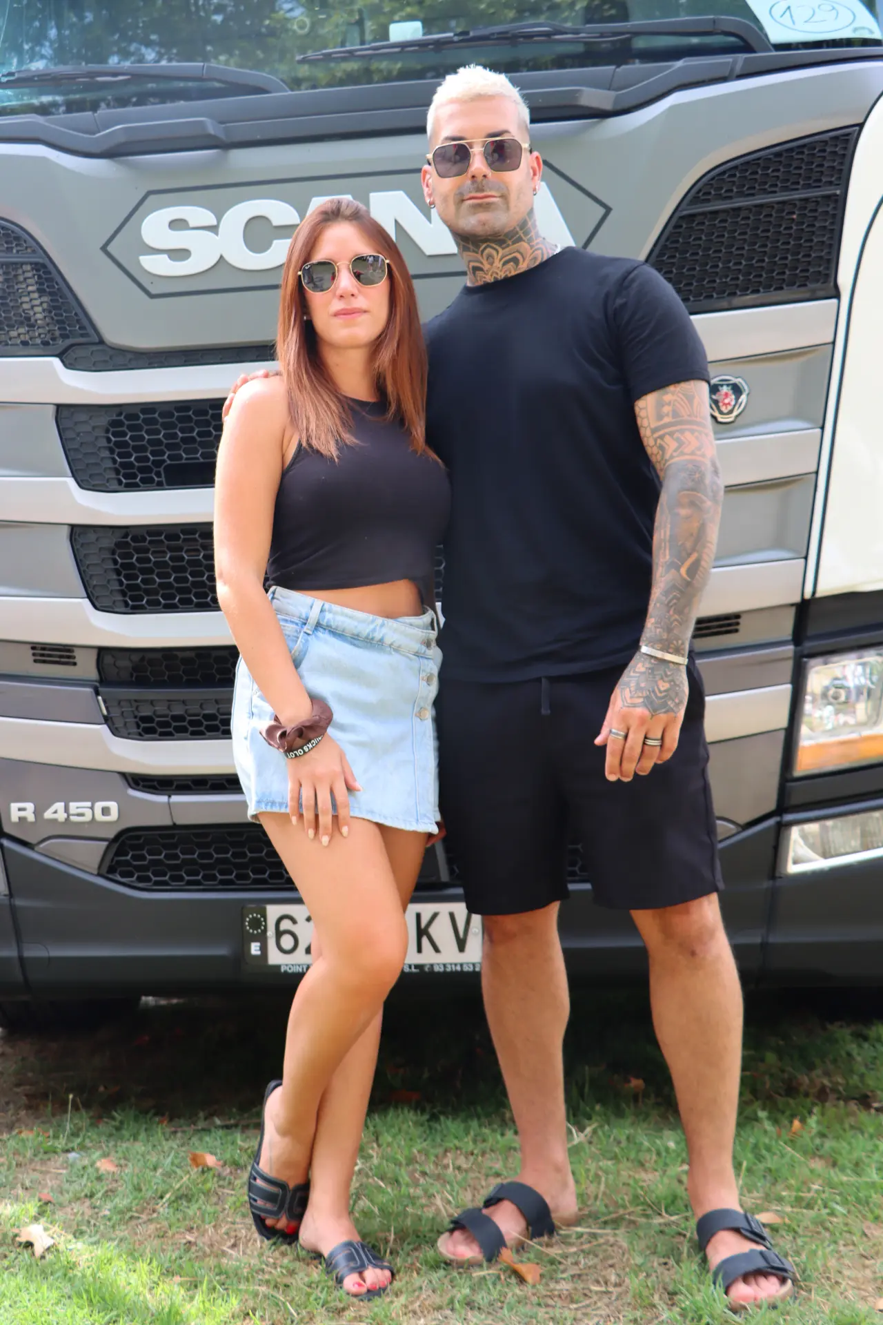 Dos personas vestidas con ropa informal están de pie sobre la hierba delante de un gran camión Scania, utilizando un borrador automático para revisar sus notas.