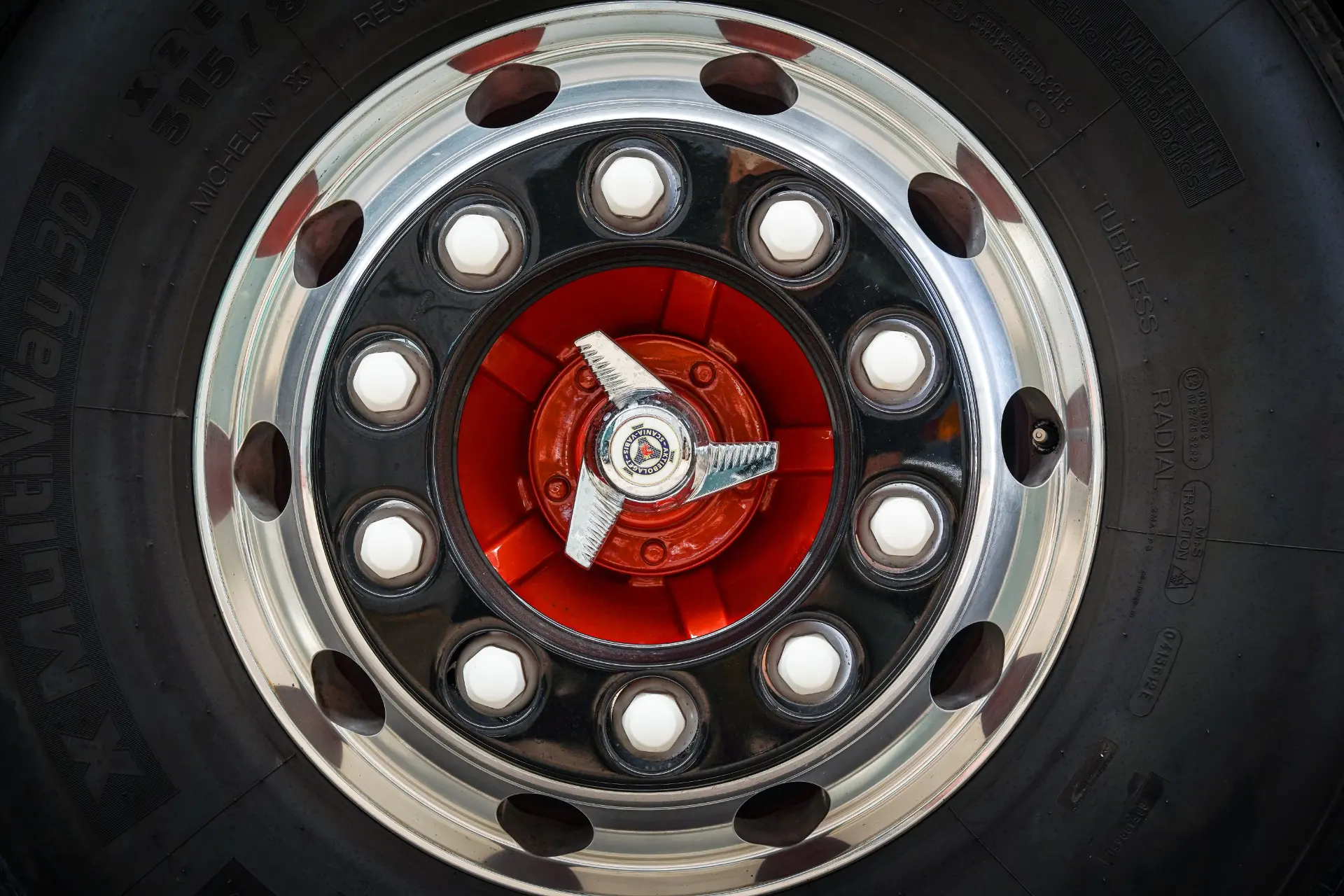 Primer plano de una llanta de camión cromada brillante con un centro rojo y múltiples tuercas de orejeta, resaltada en el reflejo de la superficie pulida como si hubiera sido procesada por un Borrador automático.