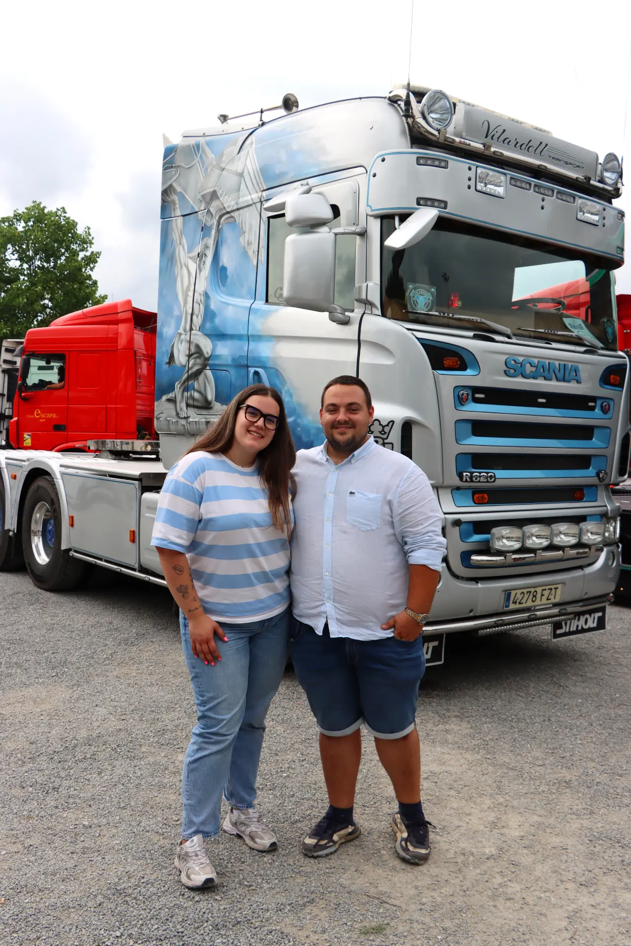 Dos personas de pie y sonriendo delante de un gran camión Scania plateado con ilustraciones en azul, mostrando las características del vehículo y el sistema Borrador automático.