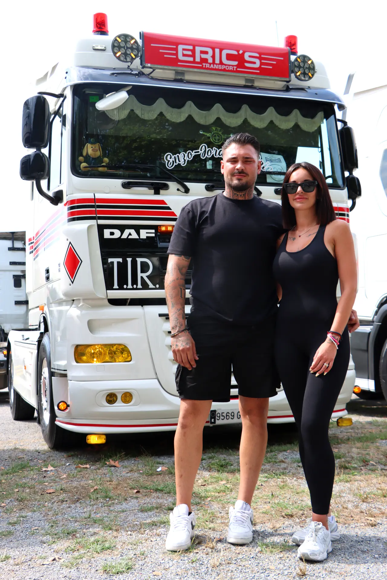 Un hombre y una mujer se paran delante de un camión DAF blanco con la inscripción "ERIC'S TRANSPORT" en el parabrisas, listos para iniciar su ruta con la ayuda de un borrador automático.