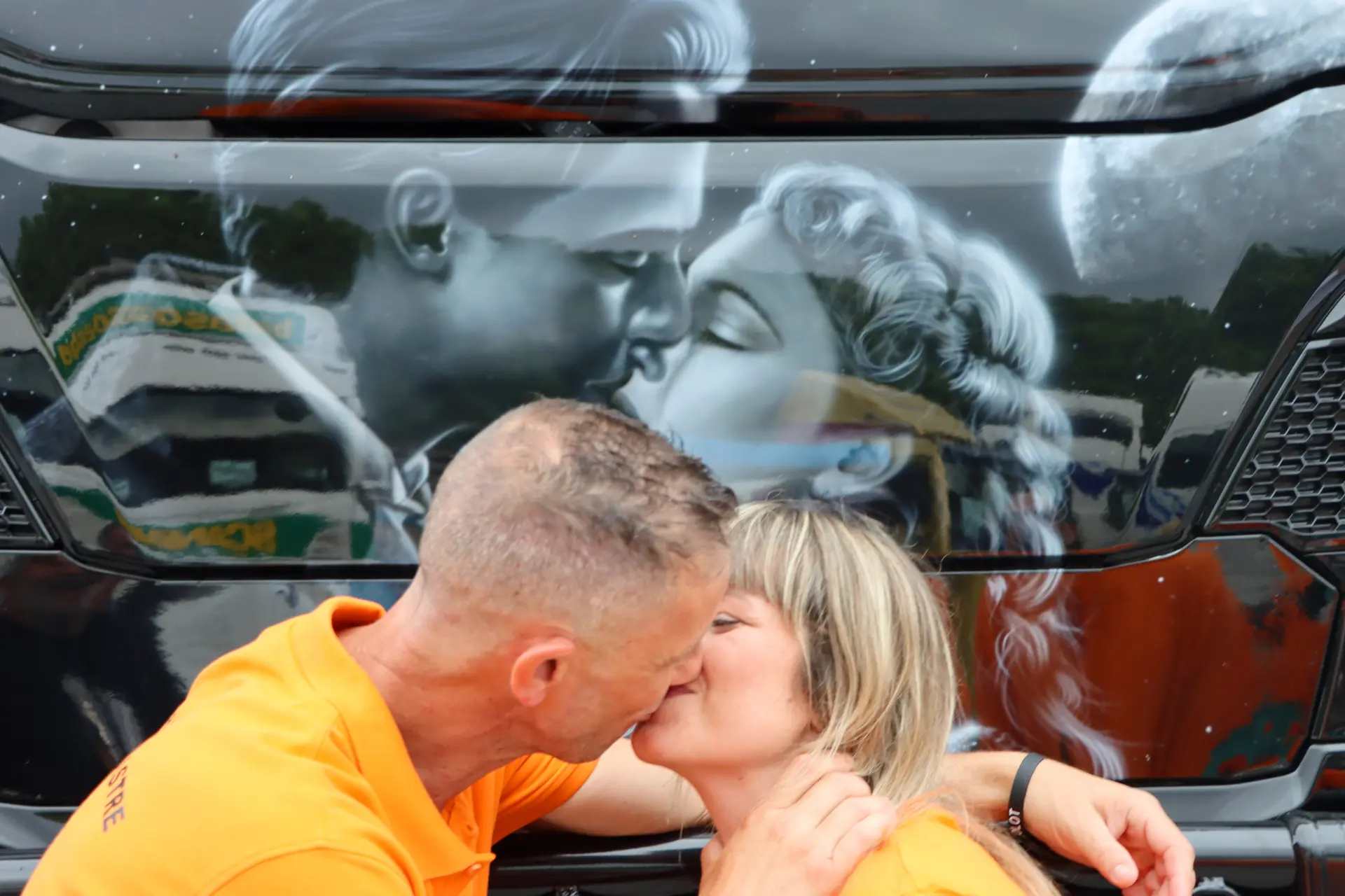 Una pareja con camisas naranjas se besa delante de un camión pintado con una escena romántica en blanco y negro en la XI Concentración de camiones de Olot.