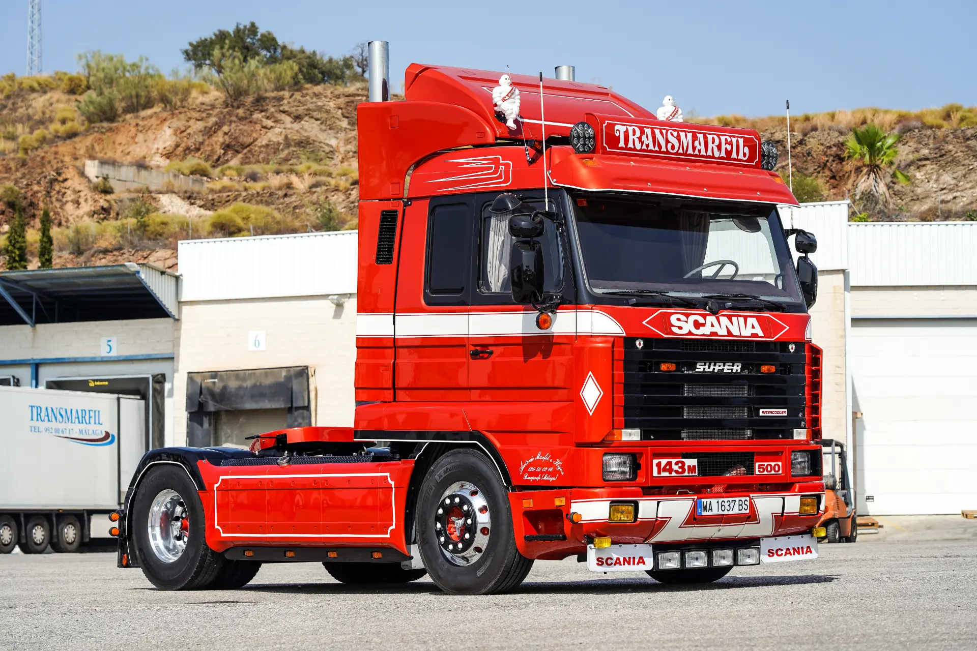 Cabina de camión Scania roja con "Transmarfil" en la parte superior delantera, equipada con un borrador automático, aparcada al aire libre cerca de un almacén.