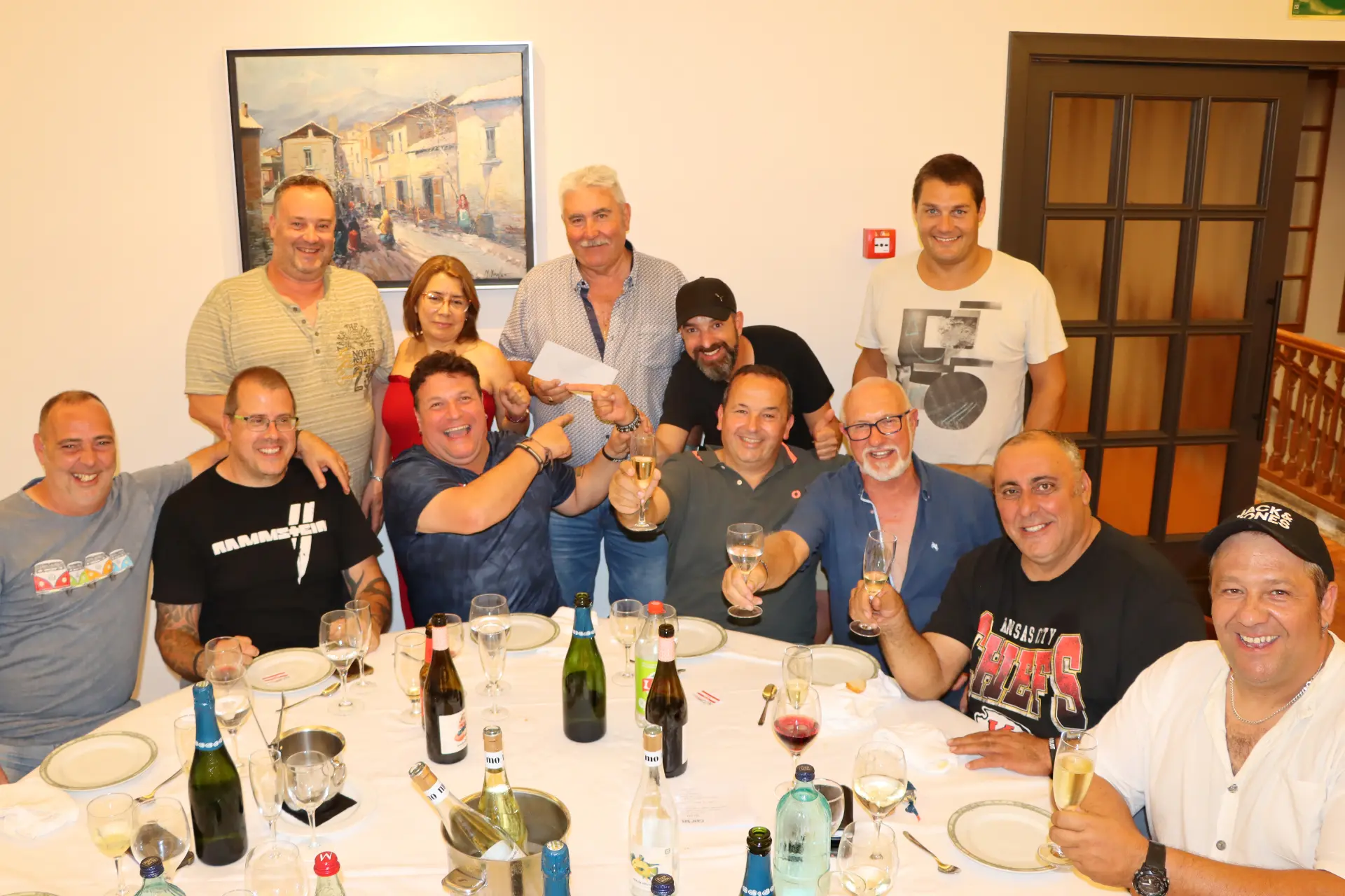 Un grupo de personas sonriendo y levantando copas alrededor de una mesa de comedor con bebidas y platos vacíos.