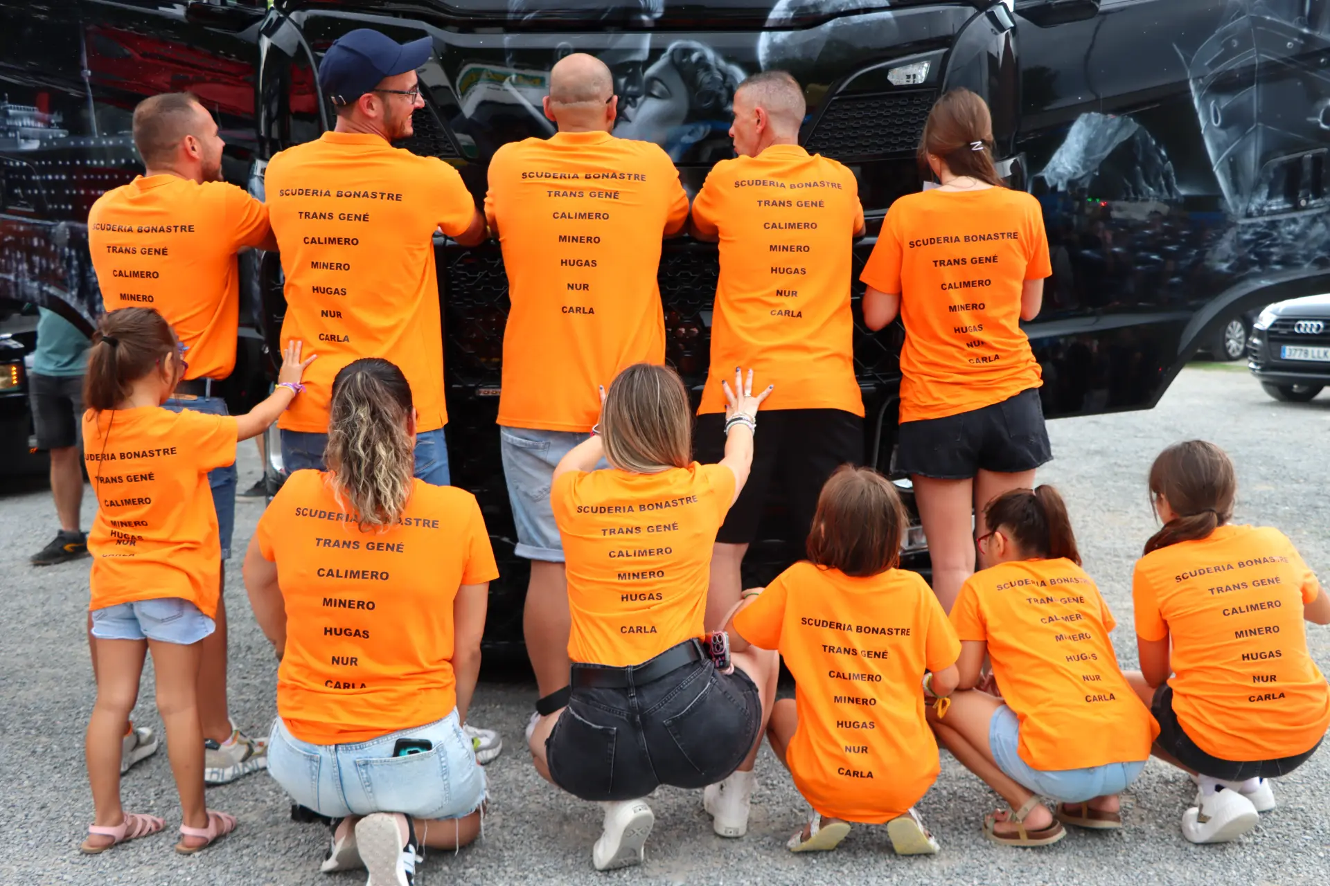 Un grupo con camisetas naranjas a juego se arrodilla y permanece de espaldas a la cámara al aire libre, probablemente participando en la XI Concentración de camiones de Olot.
