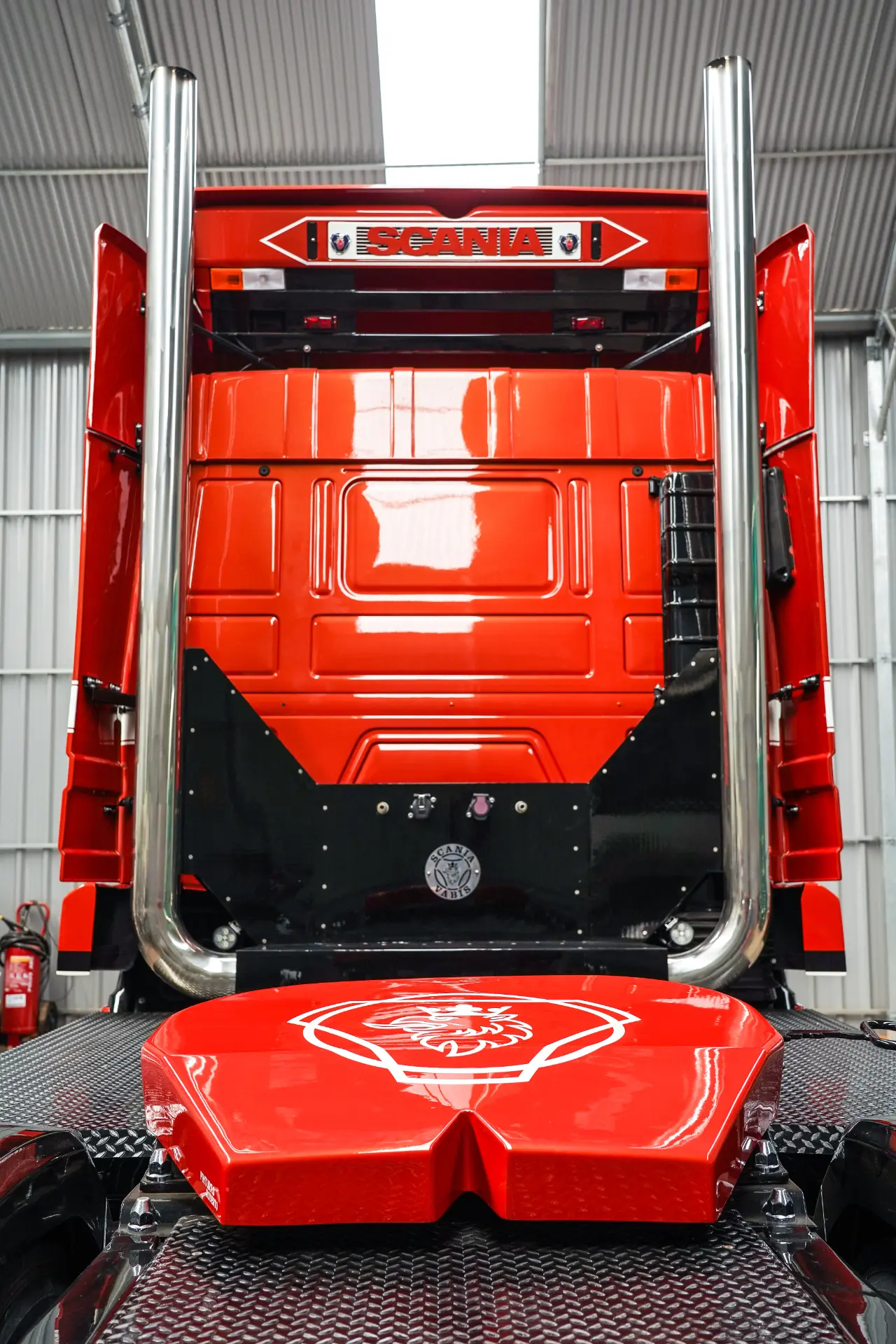 Vista trasera de la cabina de un camión Scania rojo con tubos de escape cromados y un borrador automático en un garaje.