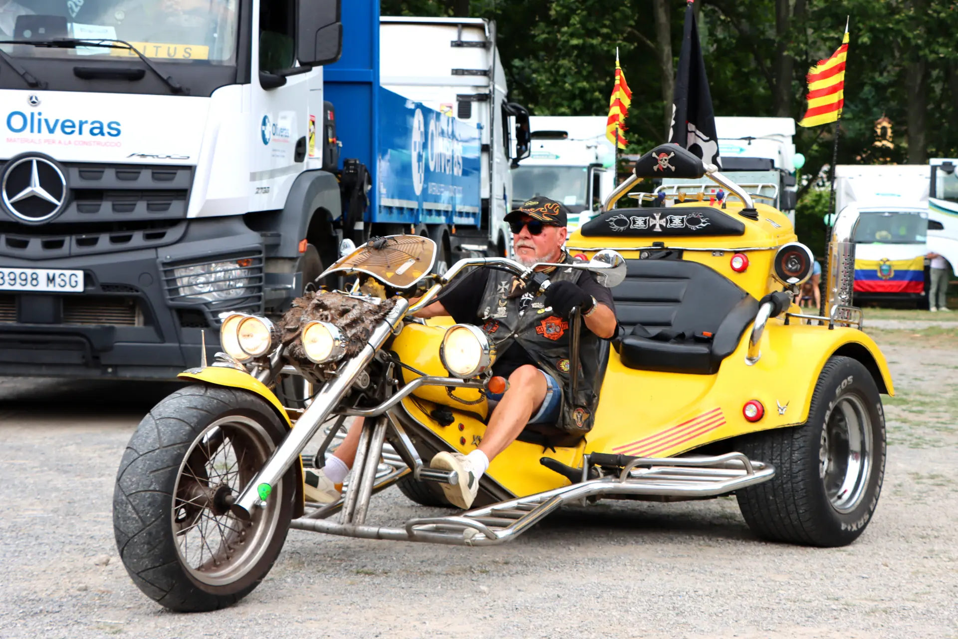 Un hombre con gafas de sol conduce una moto triciclo amarilla personalizada en un aparcamiento durante la concentración de camiones de Olot, con camiones alineados al fondo.
