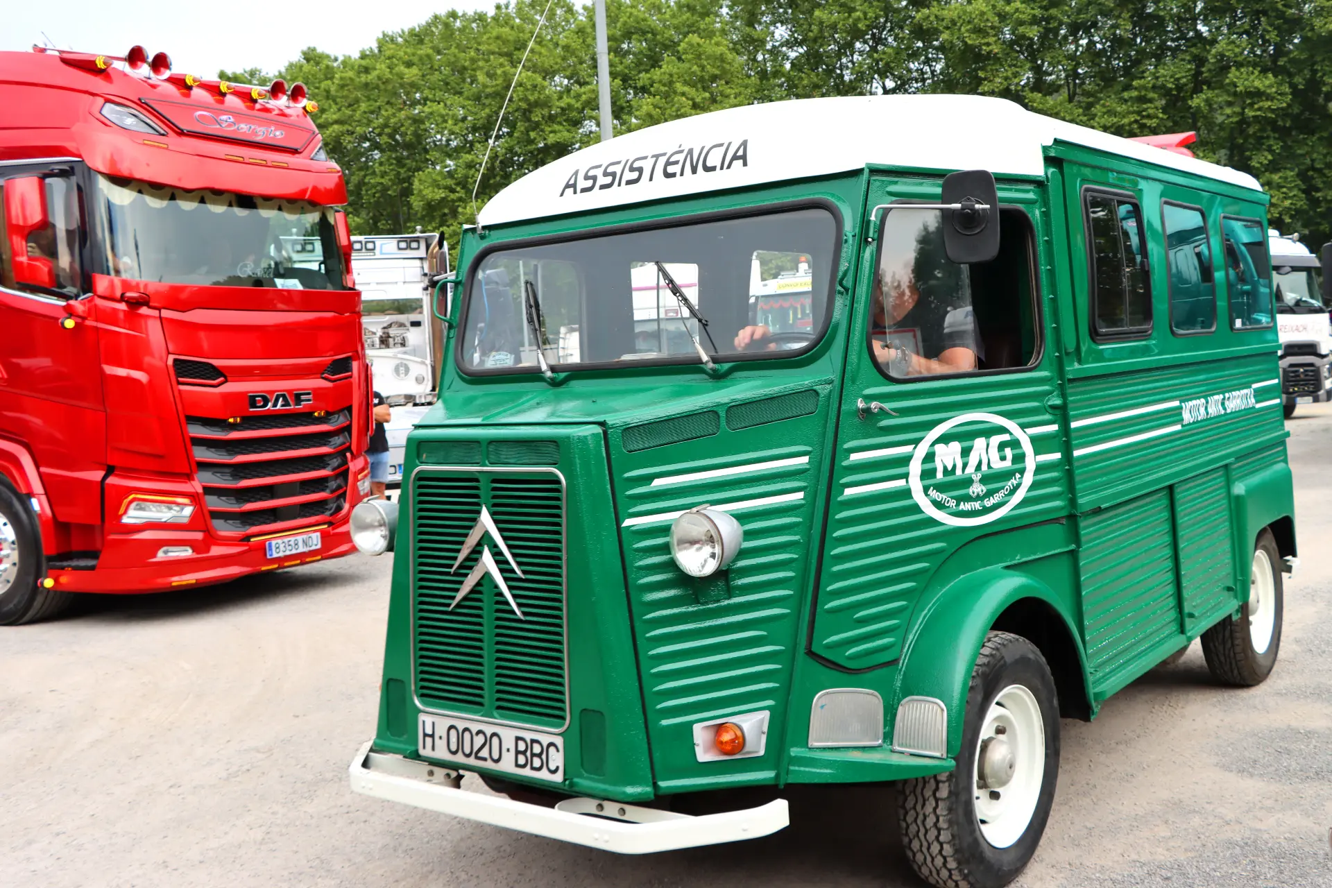 Una furgoneta Citroën verde de época con la inscripción "Assistência" en la parte superior está aparcada cerca de un camión rojo en una concentración de camiones al aire libre en Olot.