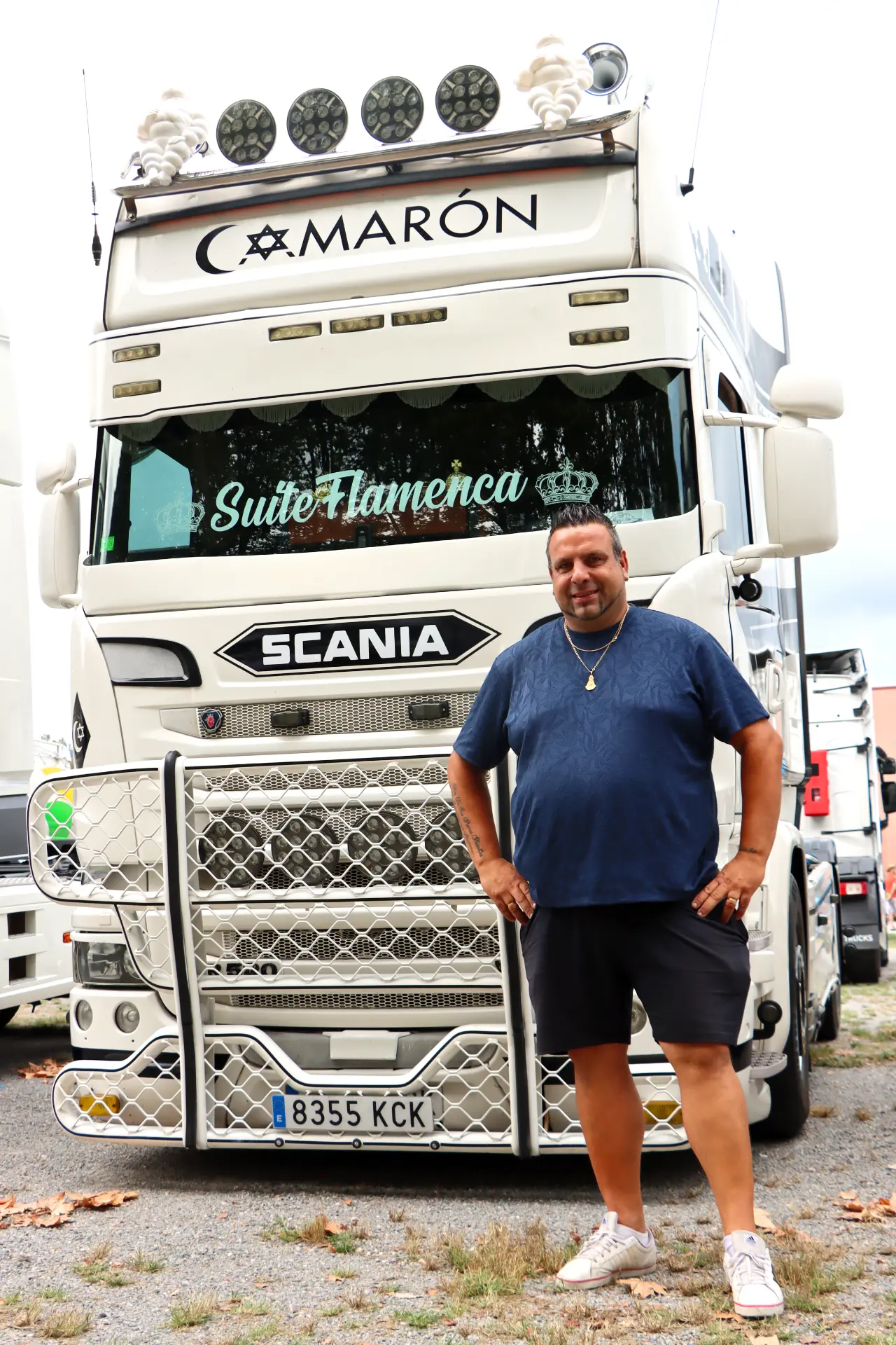 Un hombre en pantalones cortos sonríe delante de un camión Scania blanco decorado con la etiqueta "Suite Flamenca" en la XI Concentración de camiones de Olot.