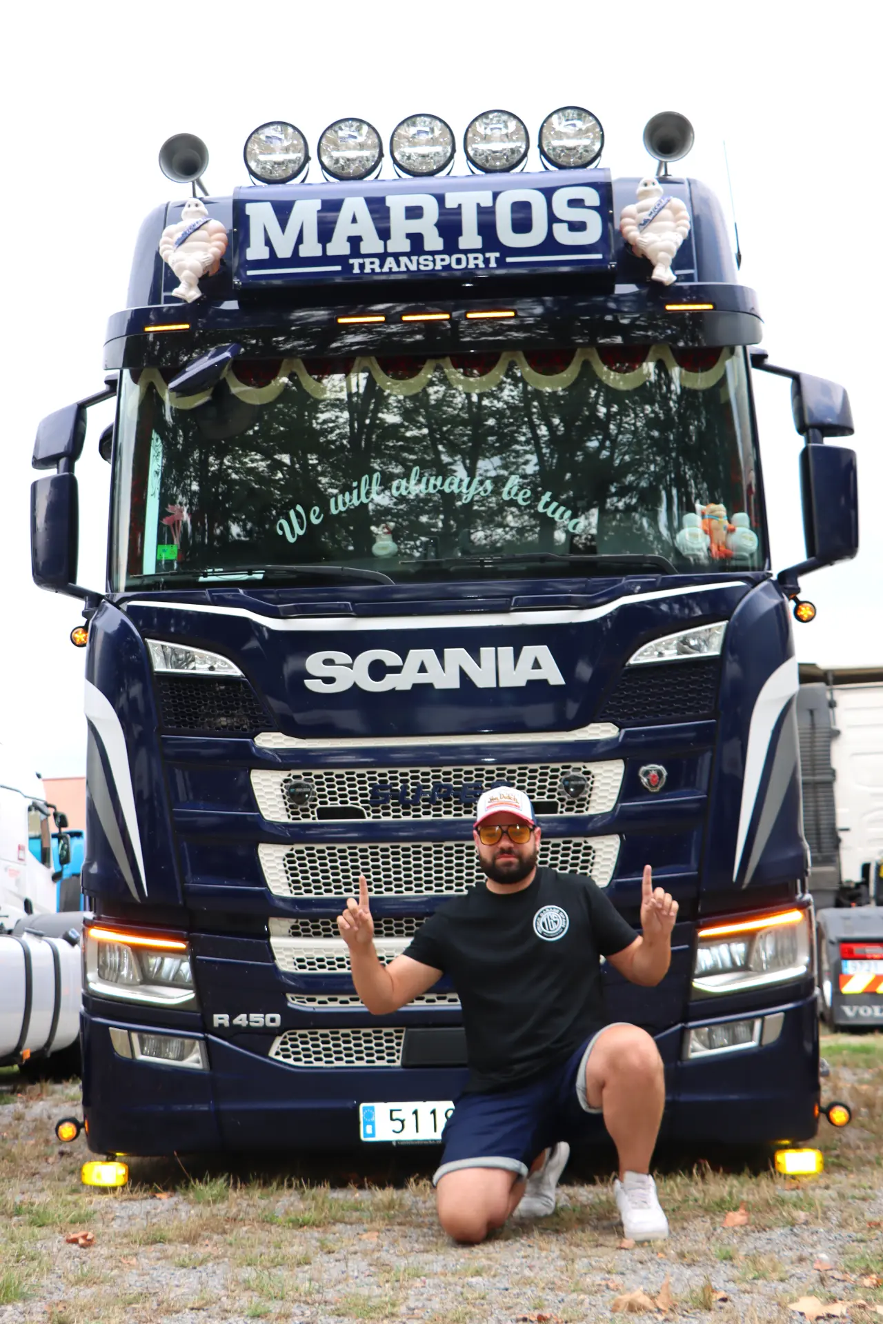 Un hombre se arrodilla y señala hacia arriba delante de un gran camión Scania azul con la etiqueta "Martos Transport" en una concentración de camiones en Olot.