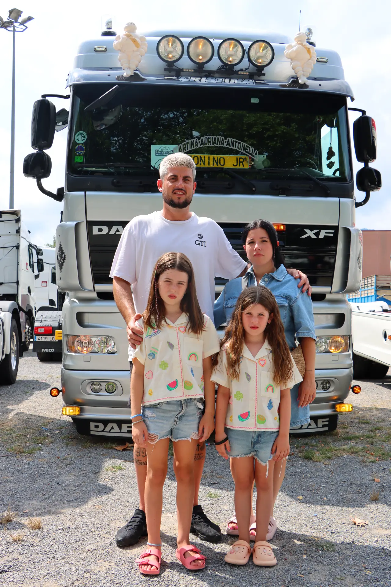 Una familia de cuatro miembros posa delante de un gran camión plateado en la XI Concentración de camiones de Olot en un aparcamiento en un día soleado.