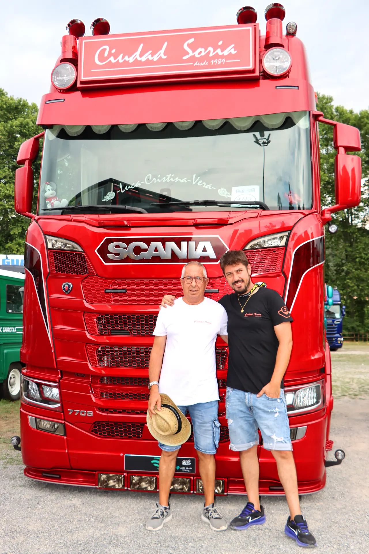 Dos hombres sonríen delante de un gran camión Scania rojo con la inscripción "Ciudad Soria" en la XI Concentración de camiones de Olot.
